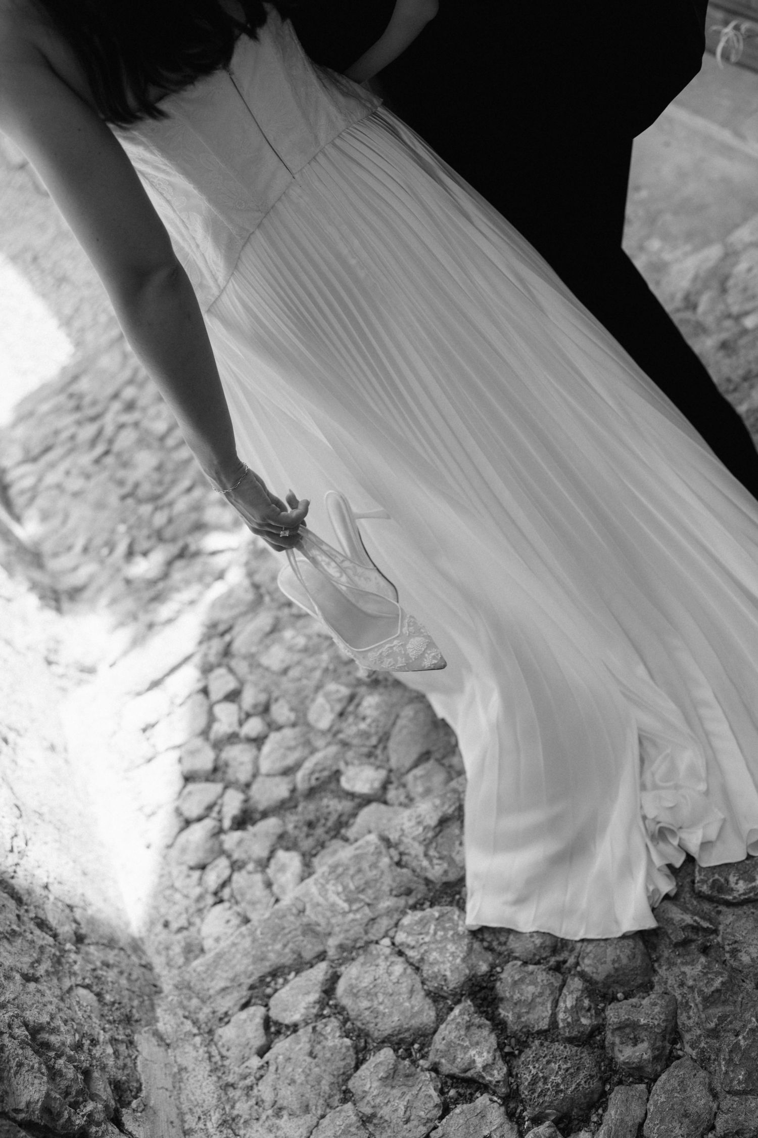 Black and white photo of a bride in a flowing pleated wedding gown walking over sunlit cobblestones, holding the dress with one hand.