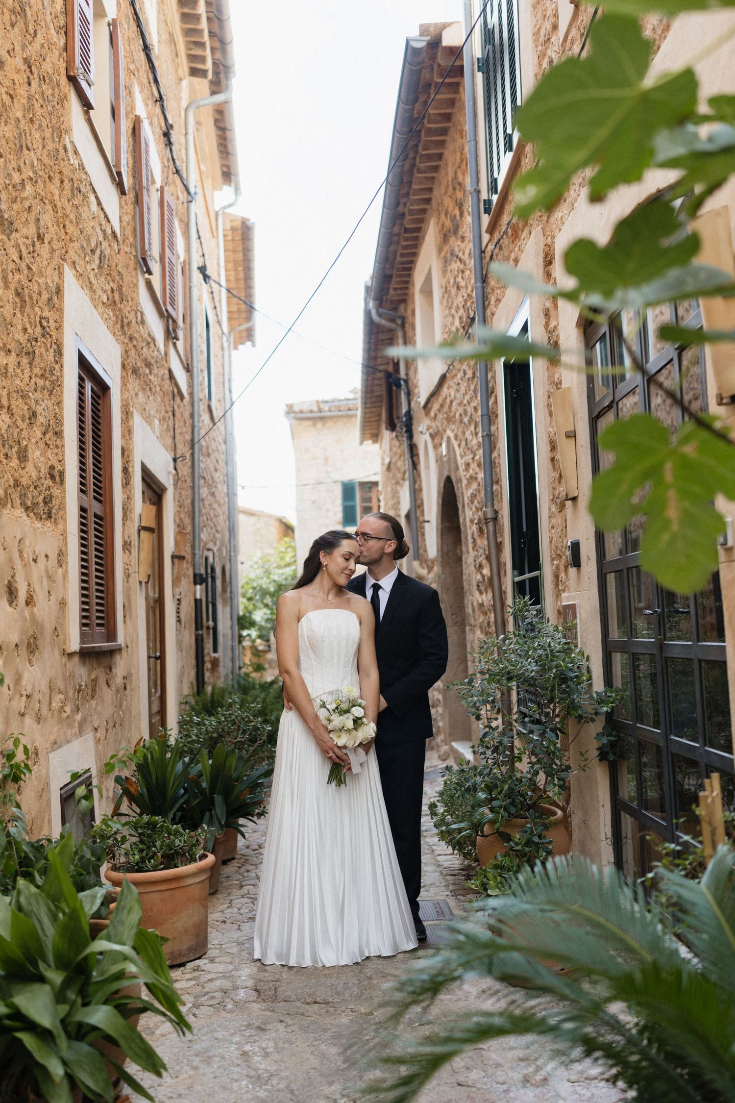 Bride in a white gown holding a bouquet embraces a groom in a dark suit in a narrow stone alley lined with potted plants.