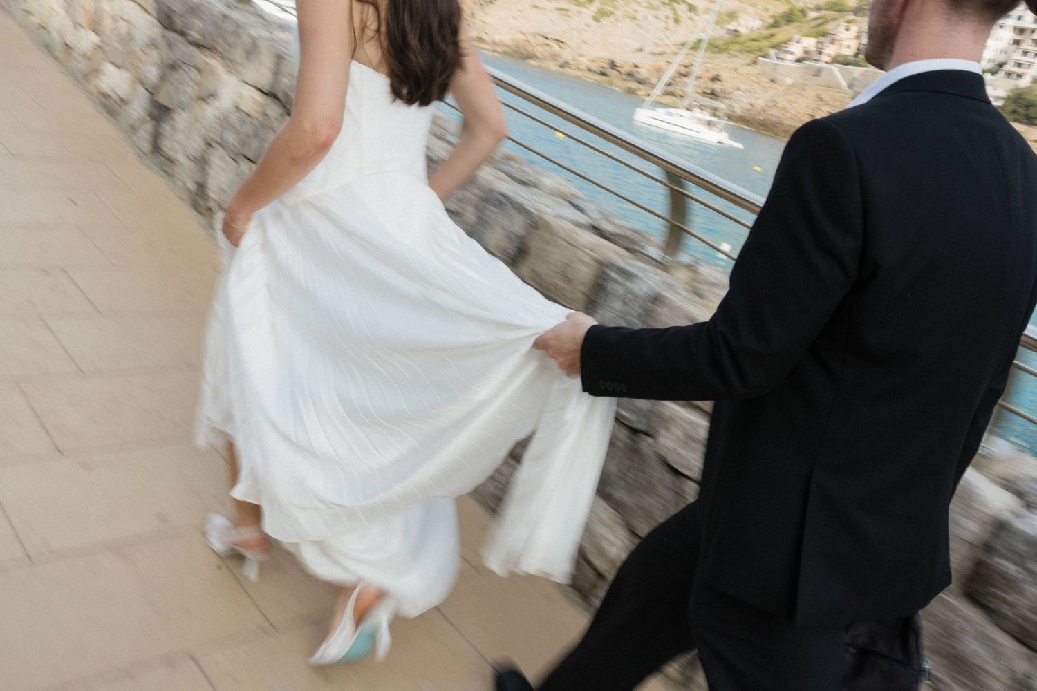 Bride in a white wedding gown and blue heels walks along a stone seaside path as the groom in a black suit holds her dress.