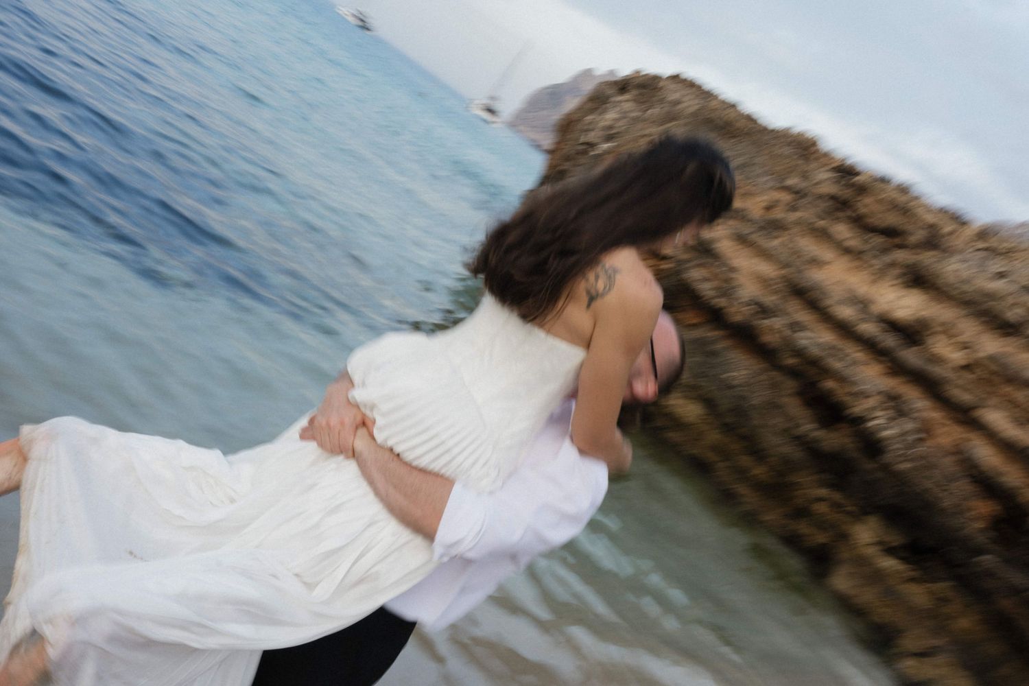 Bride in a flowing white dress embraces her partner in a romantic dip on a rocky beach beside the calm blue sea.