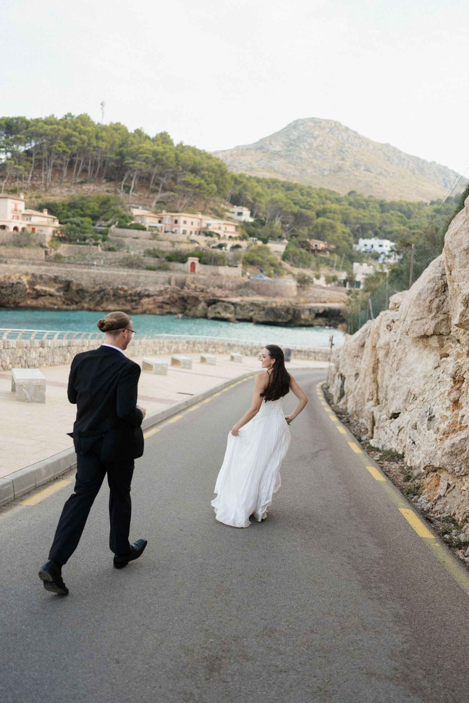 Bride in a flowing white gown and groom in a black suit walk along a coastal road beside rocky cliffs, turquoise sea and a hillside village.