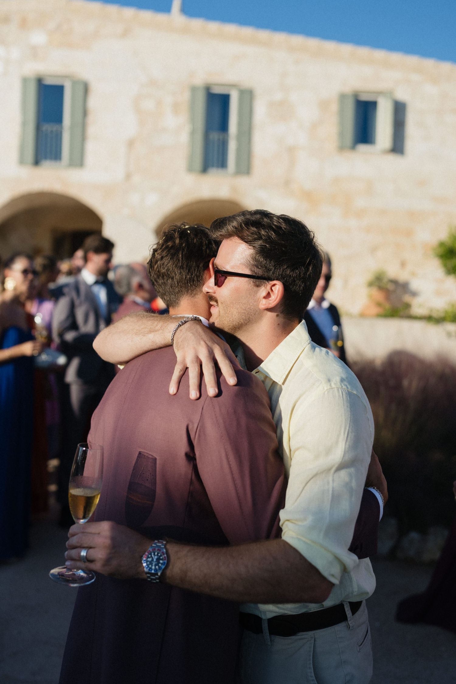 Two men in a warm embrace at an outdoor wedding celebration, one holding a champagne glass, with guests and a stone building behind them.