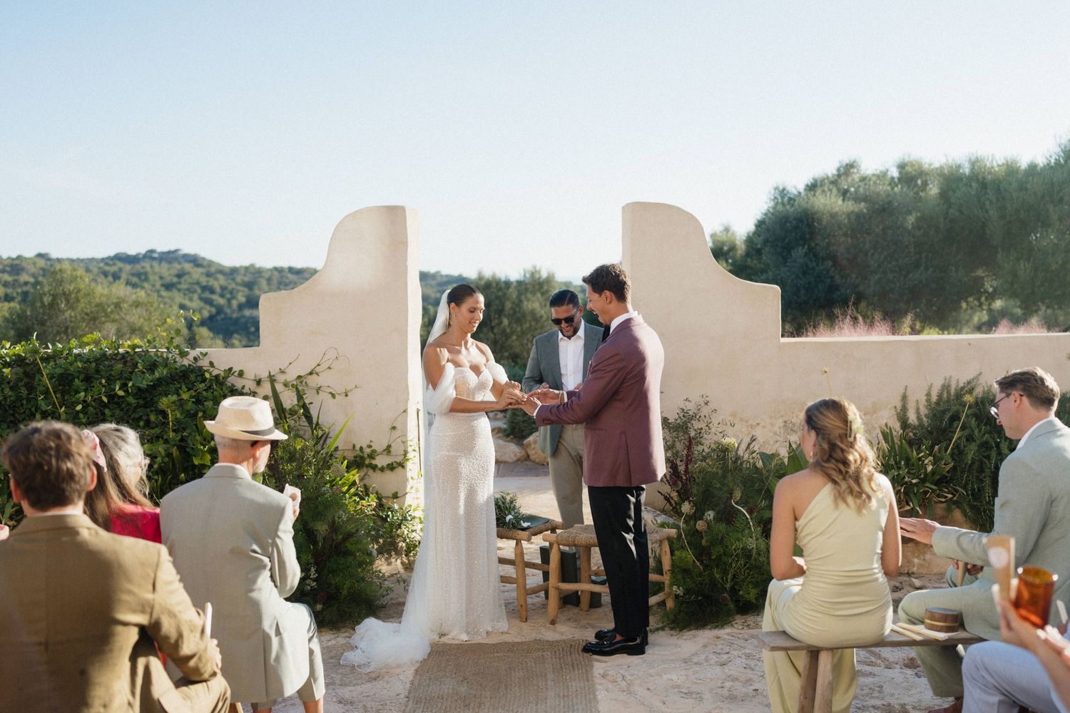 Bride in a white gown and groom in a burgundy suit exchange vows during an outdoor ceremony as seated guests watch.