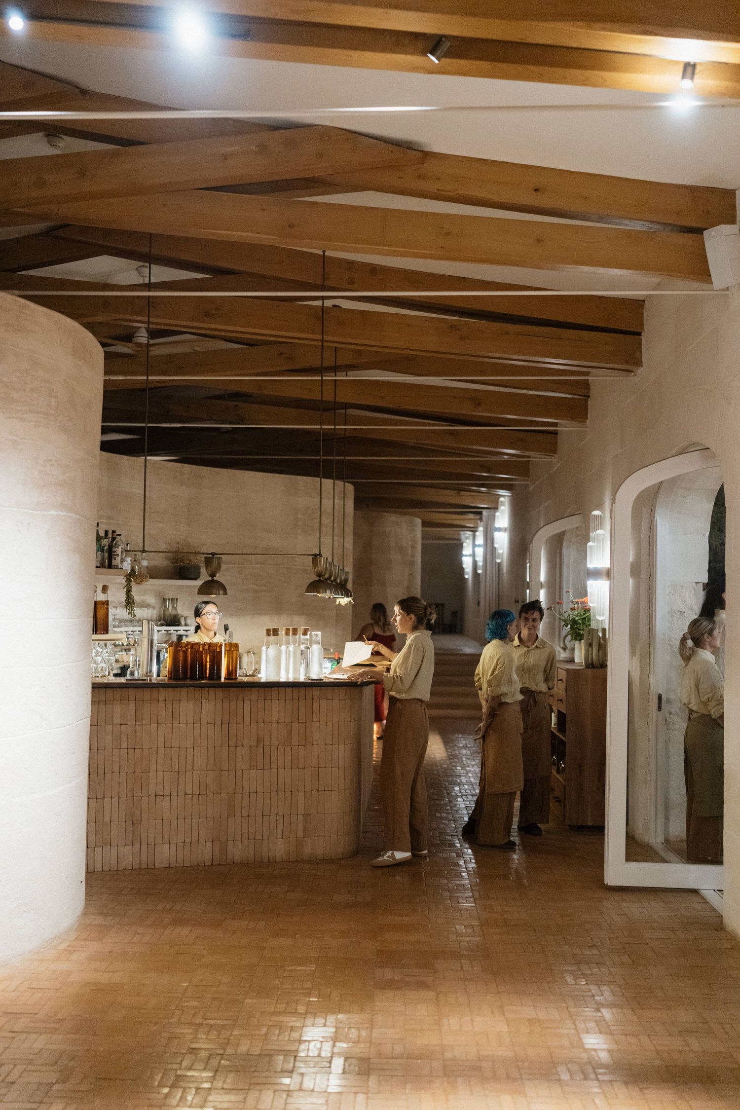 Staff in neutral uniforms work at a wooden bar inside a warm rustic venue with exposed beams and a brick floor.