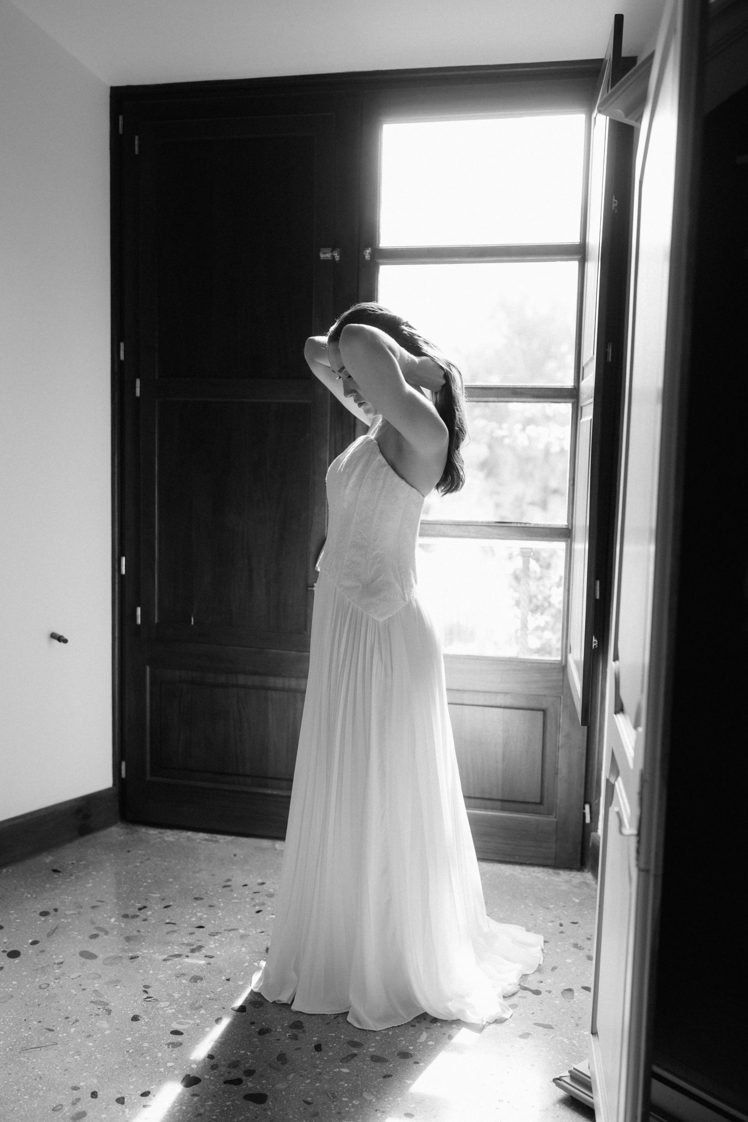 Black and white photo of a bride in a flowing white gown adjusting her hair beside a sunlit window and dark wooden doors.
