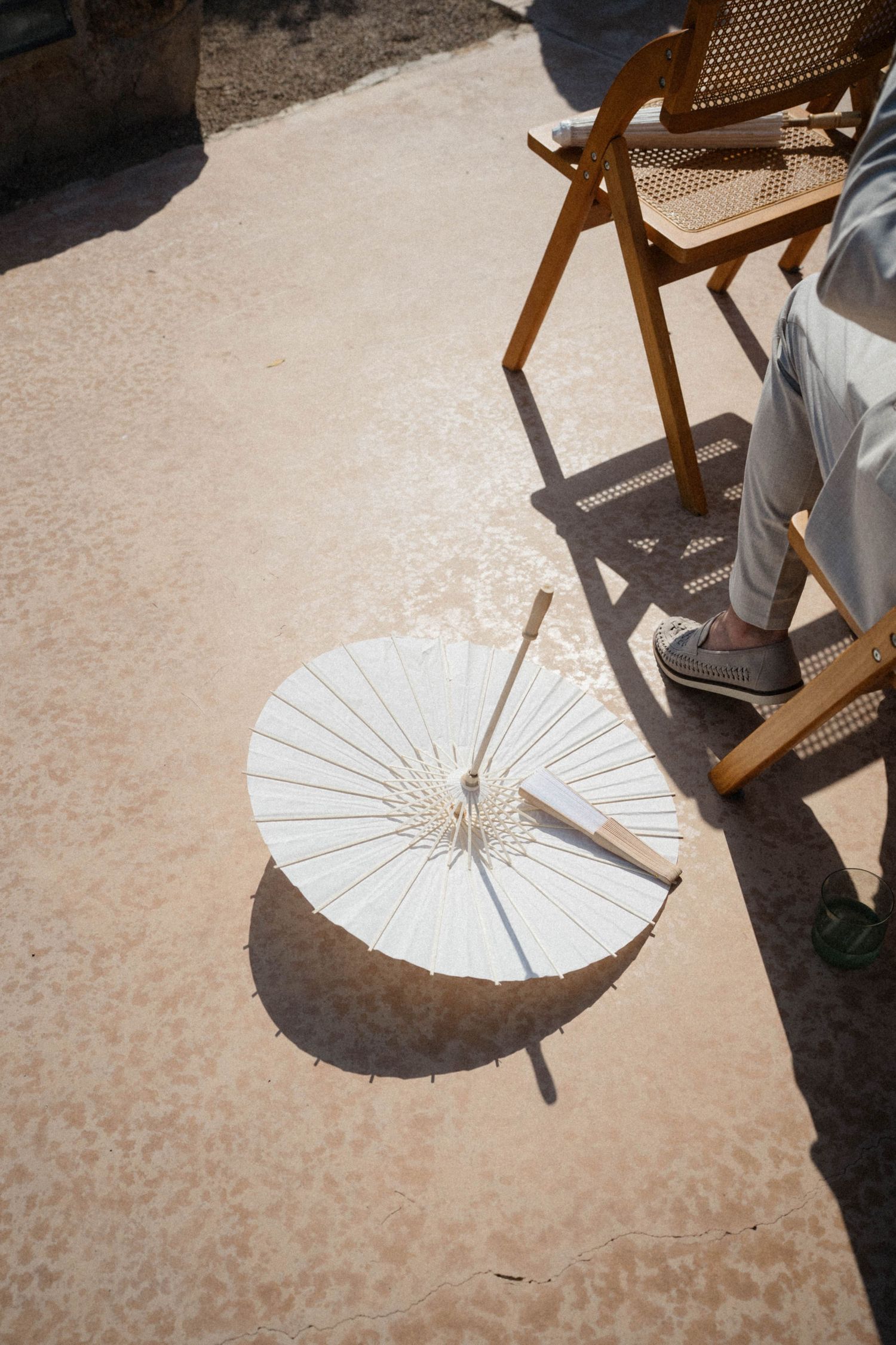 White paper parasol umbrella lying on sandy ground beside wooden chair with person's legs visible in sunlight.