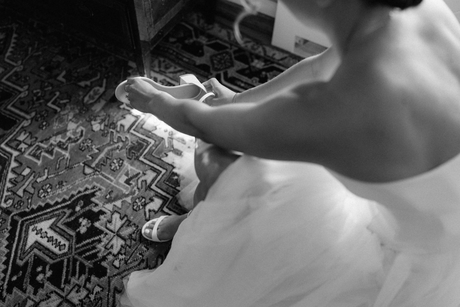 Bride in white wedding gown putting on shoes while seated on a patterned rug.