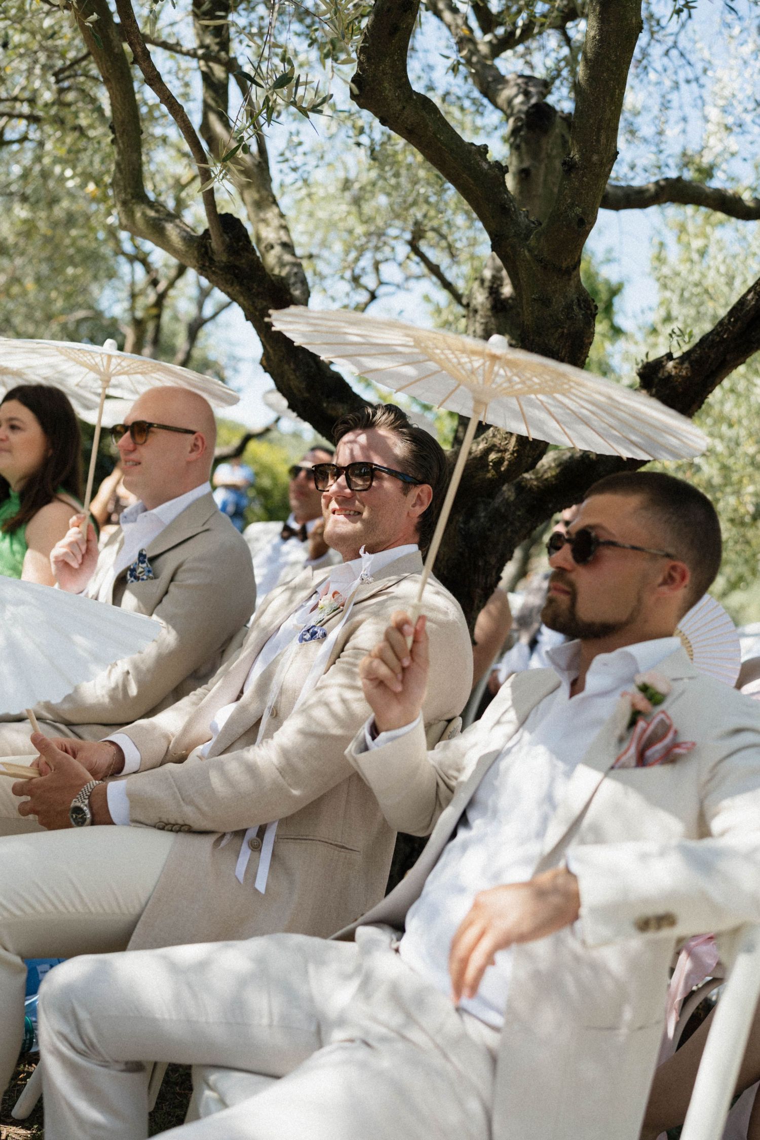 Guests in beige suits holding white parasols sit outdoors at a sunny wedding ceremony under olive trees.