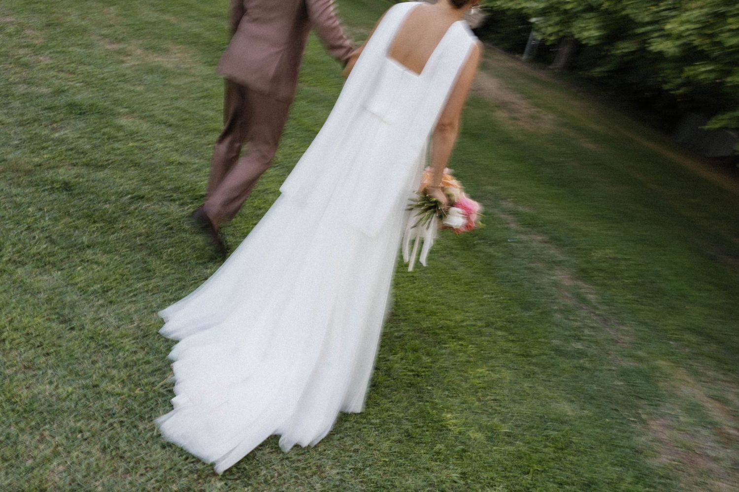 Bride in white flowing gown holding colorful bouquet walks with groom in tan suit on green grass.