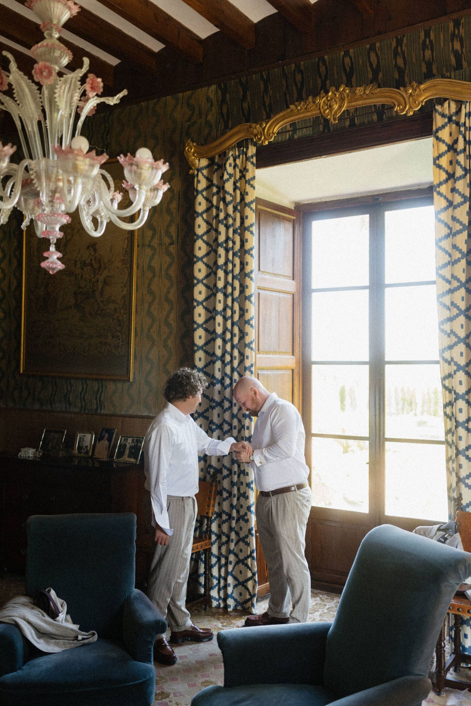 Two elderly men shaking hands in an elegant Venetian room with ornate chandelier and gold-framed window.