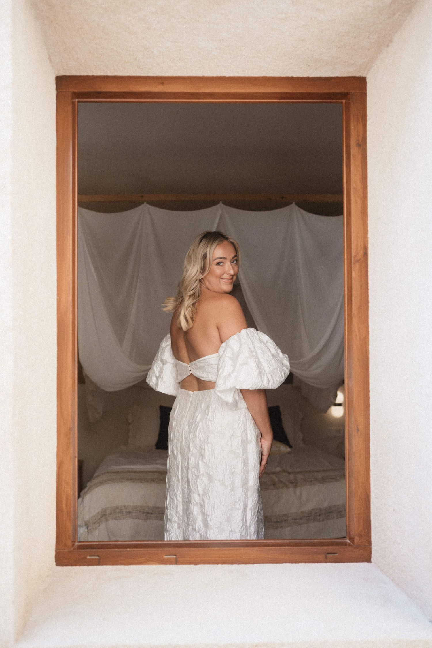 Bride in off-shoulder white textured wedding dress posing in a wooden-framed window alcove.