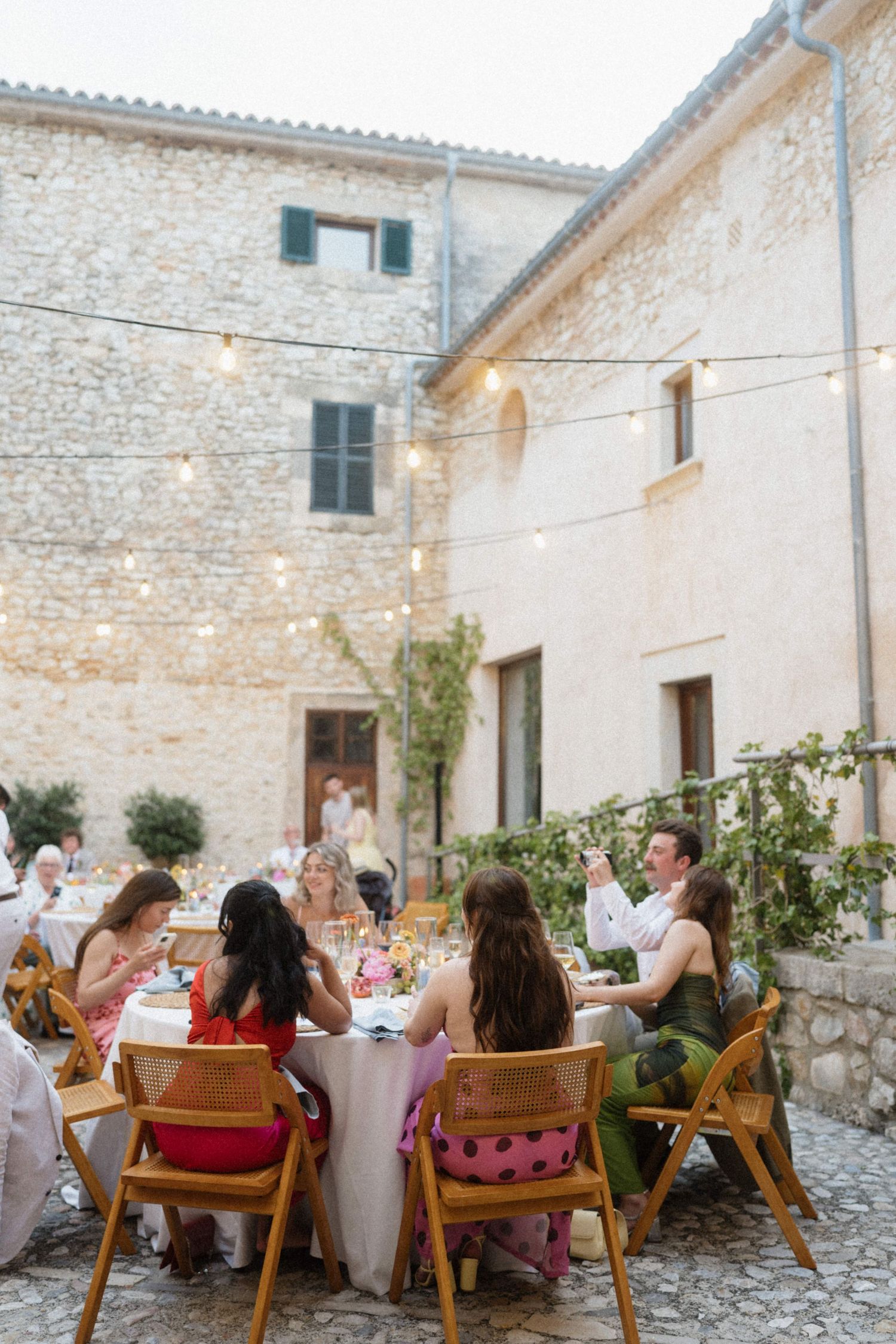 Guests dining al fresco at a rustic stone villa courtyard, illuminated by string lights at a summer evening gathering.