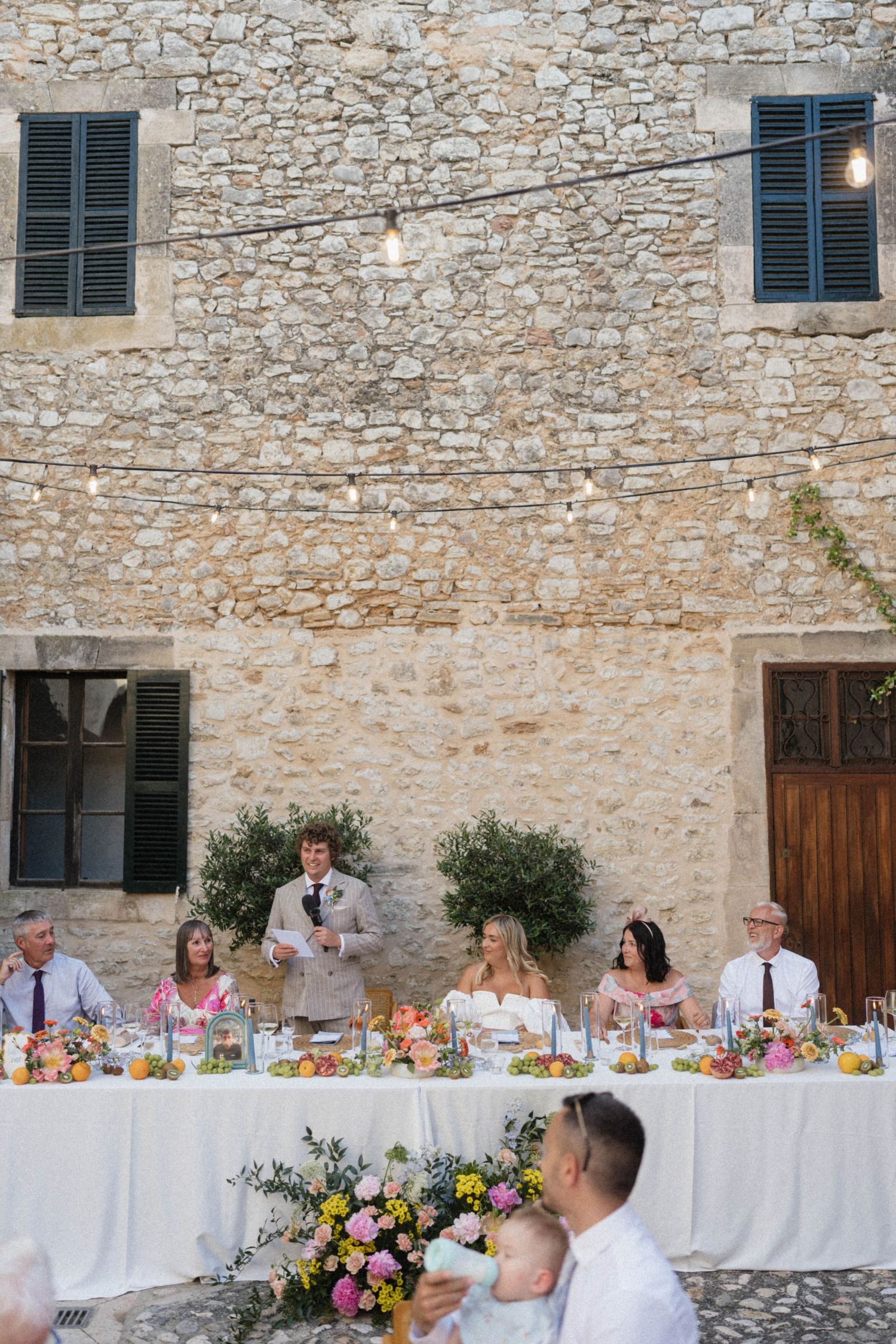 Outdoor wedding reception at a rustic stone villa with guests seated at a decorated table as the groom gives a speech.