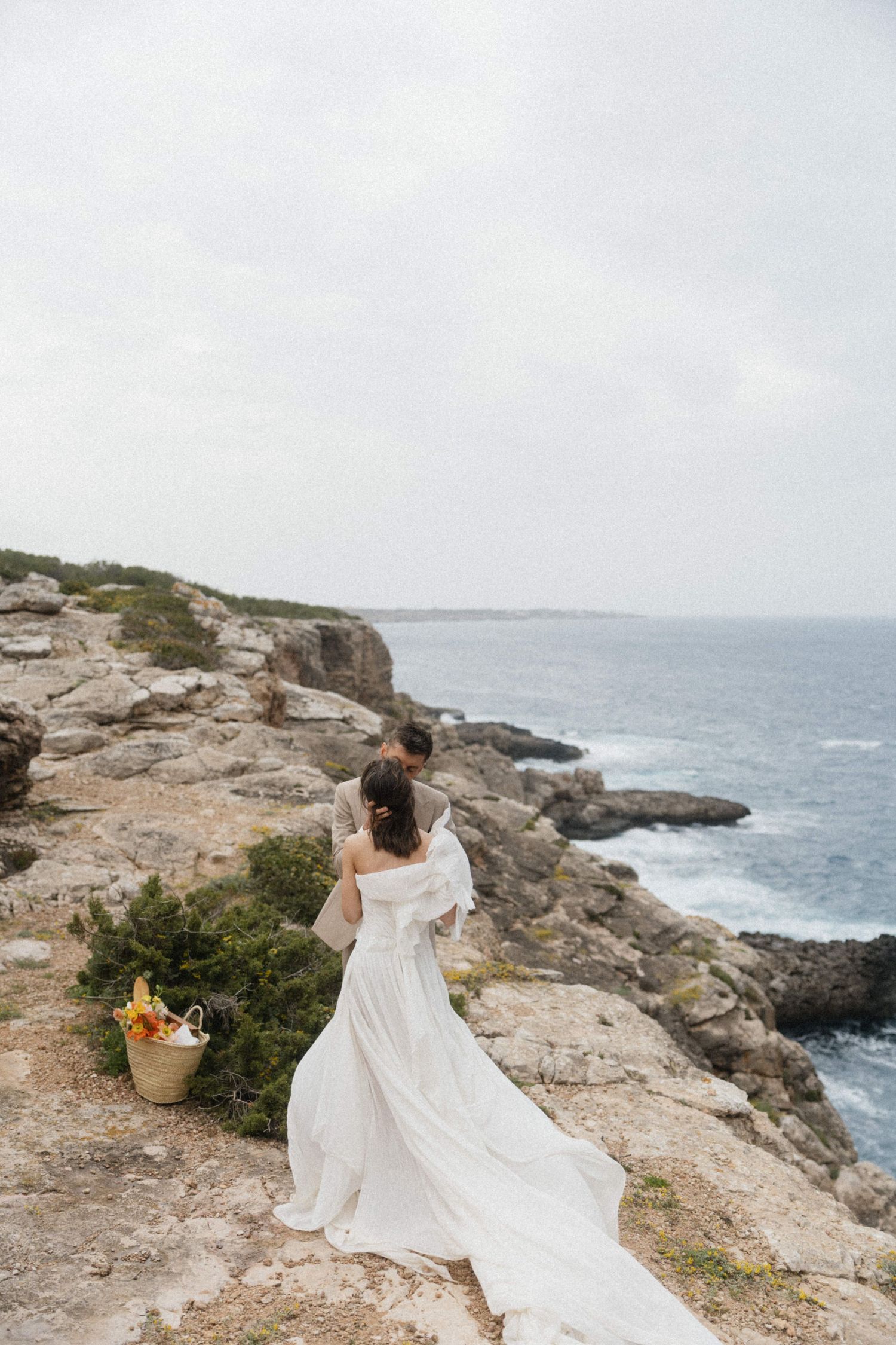 Bride and groom stand on rocky coastal cliffs overlooking the ocean, wedding dress flowing in the breeze.
