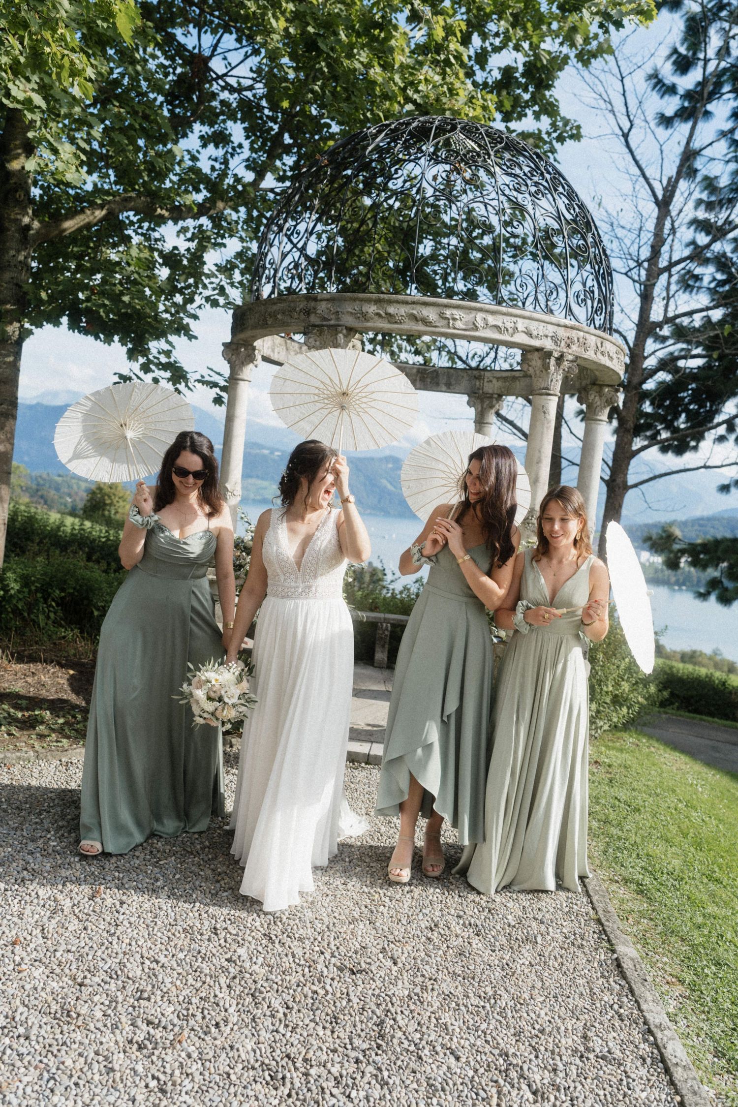 Bride and bridesmaids in sage green gowns holding white parasols under a stone gazebo with lake views.