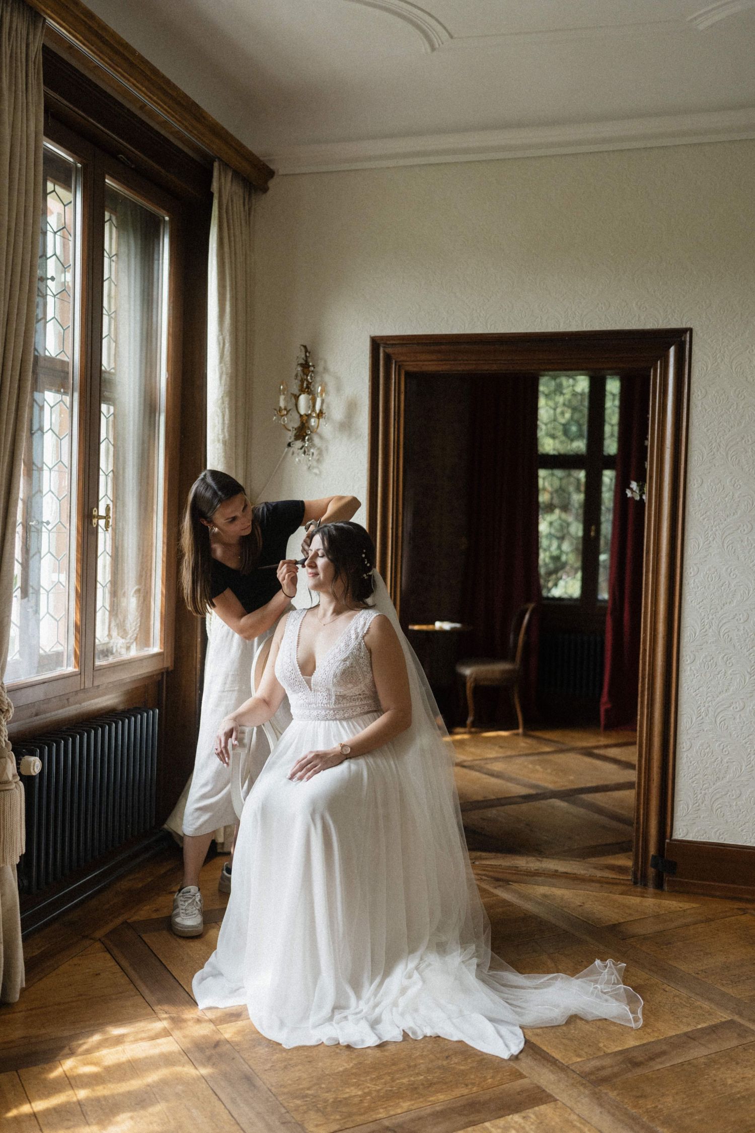 Hair stylist arranging bride's veil in a sunlit elegant room with herringbone floors on wedding day.