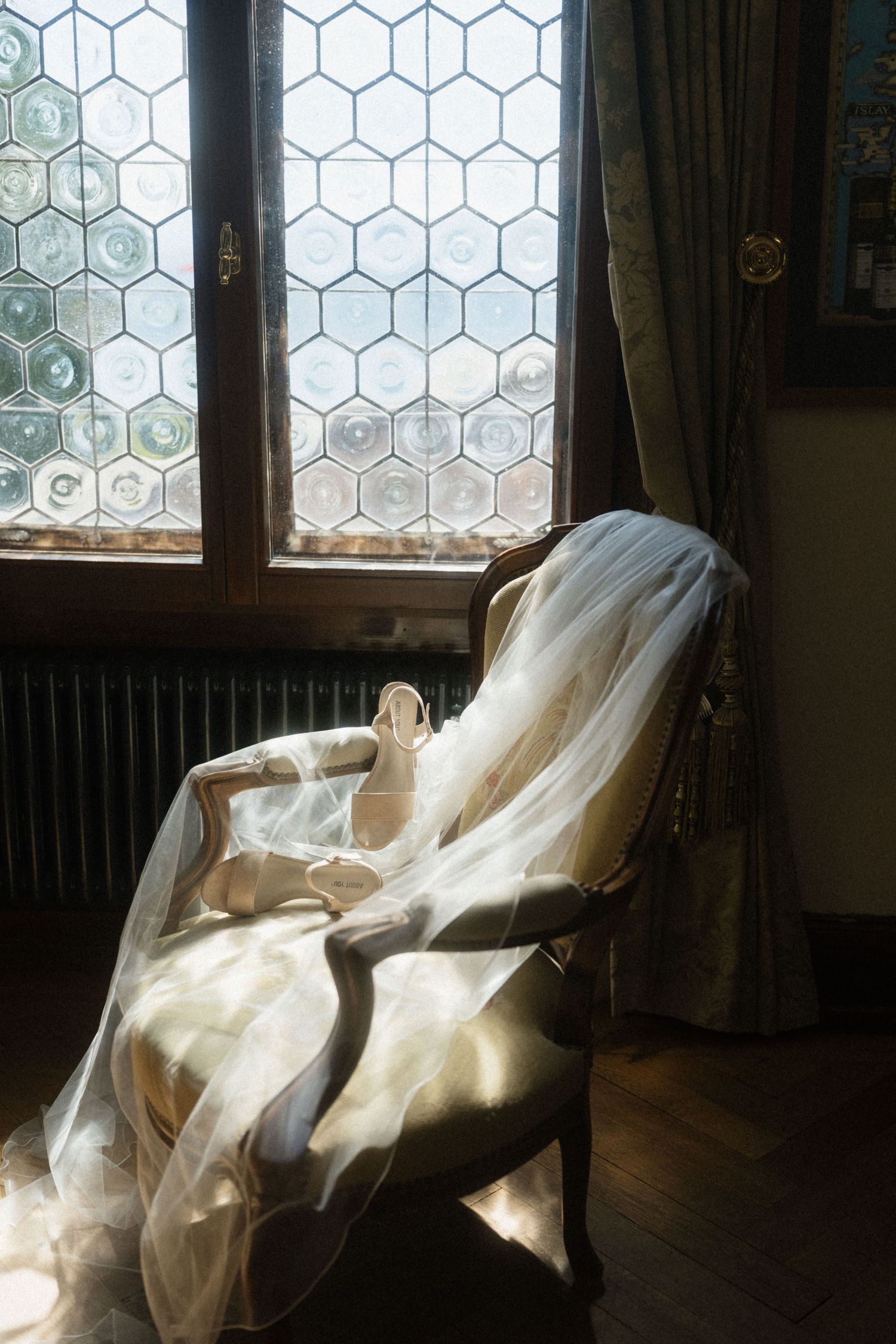 Bridal veil and wedding shoes resting on a vintage chair by a stained glass window in soft natural light.