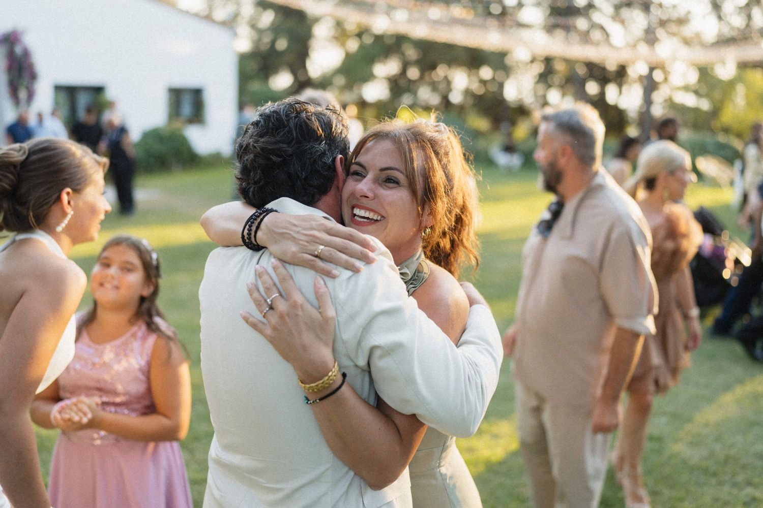 Couple embracing warmly at an outdoor wedding reception on a sunny day with guests mingling on green lawn.