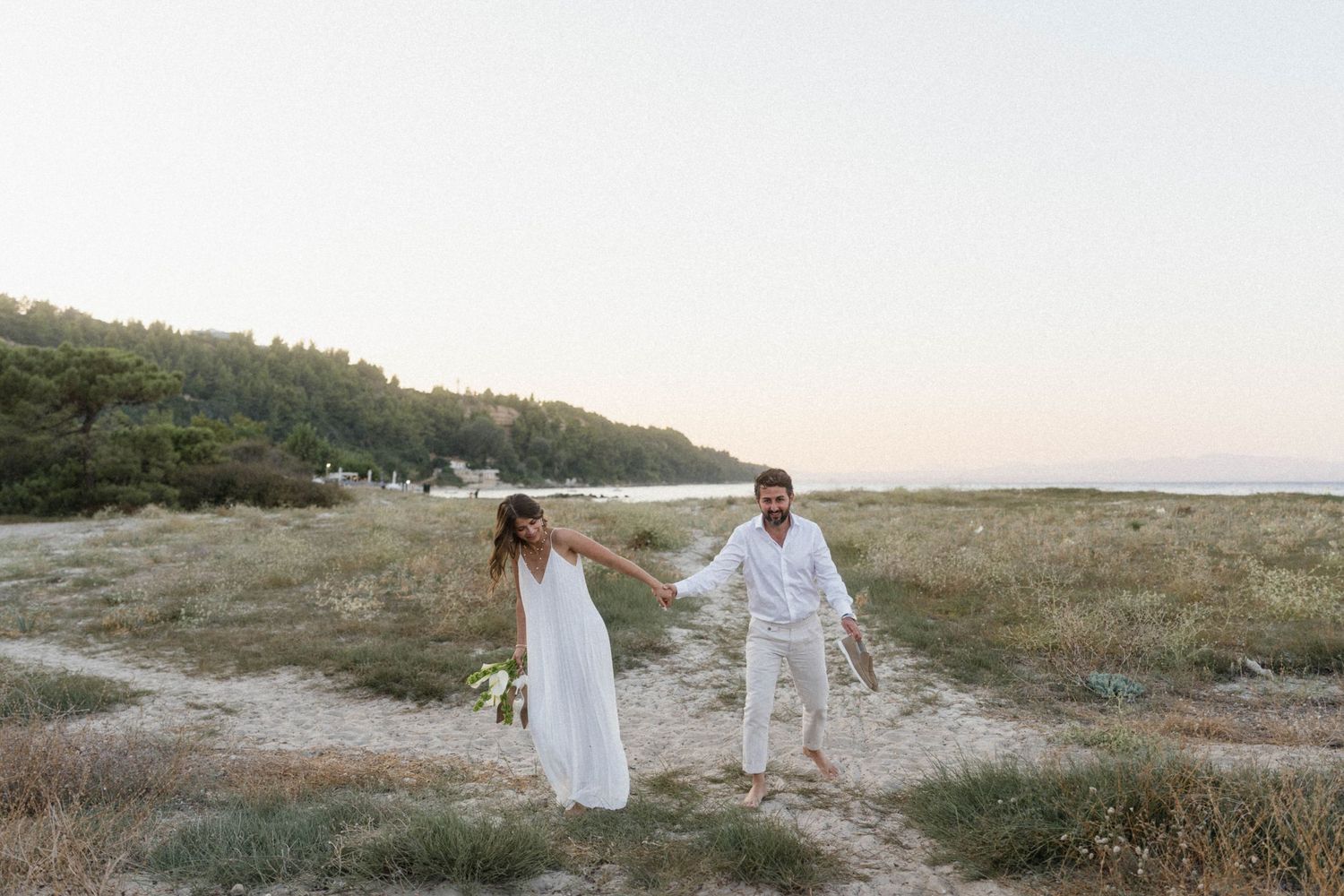 Couple in white outfits holding hands on a sandy path with green trees and a calm beach in the background.