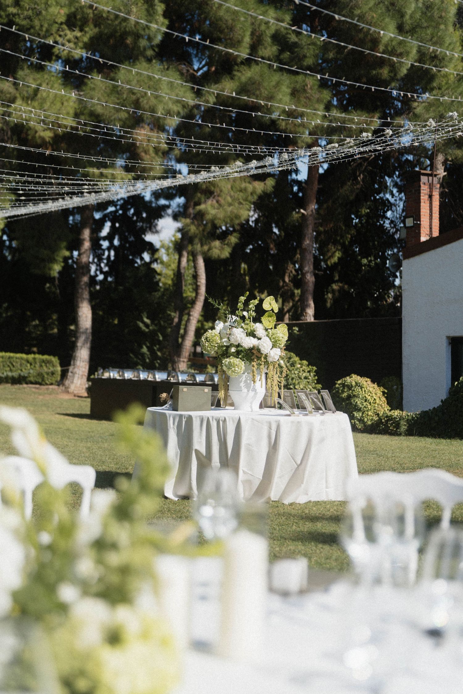 Elegant outdoor wedding reception setup with white floral centerpiece, string lights, and lush green garden backdrop.