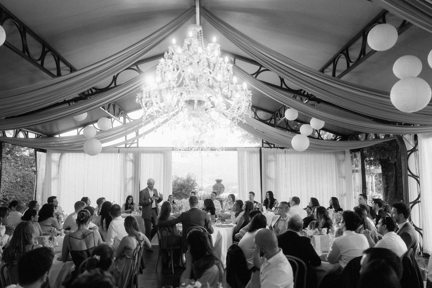 Elegant black and white wedding reception under draped tent with chandelier and globe lights above guests.