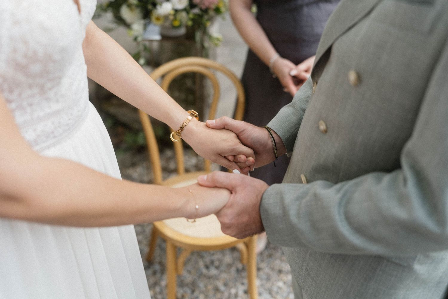 Bride and groom holding hands during outdoor wedding ceremony, bride wearing gold bracelet and white lace dress.