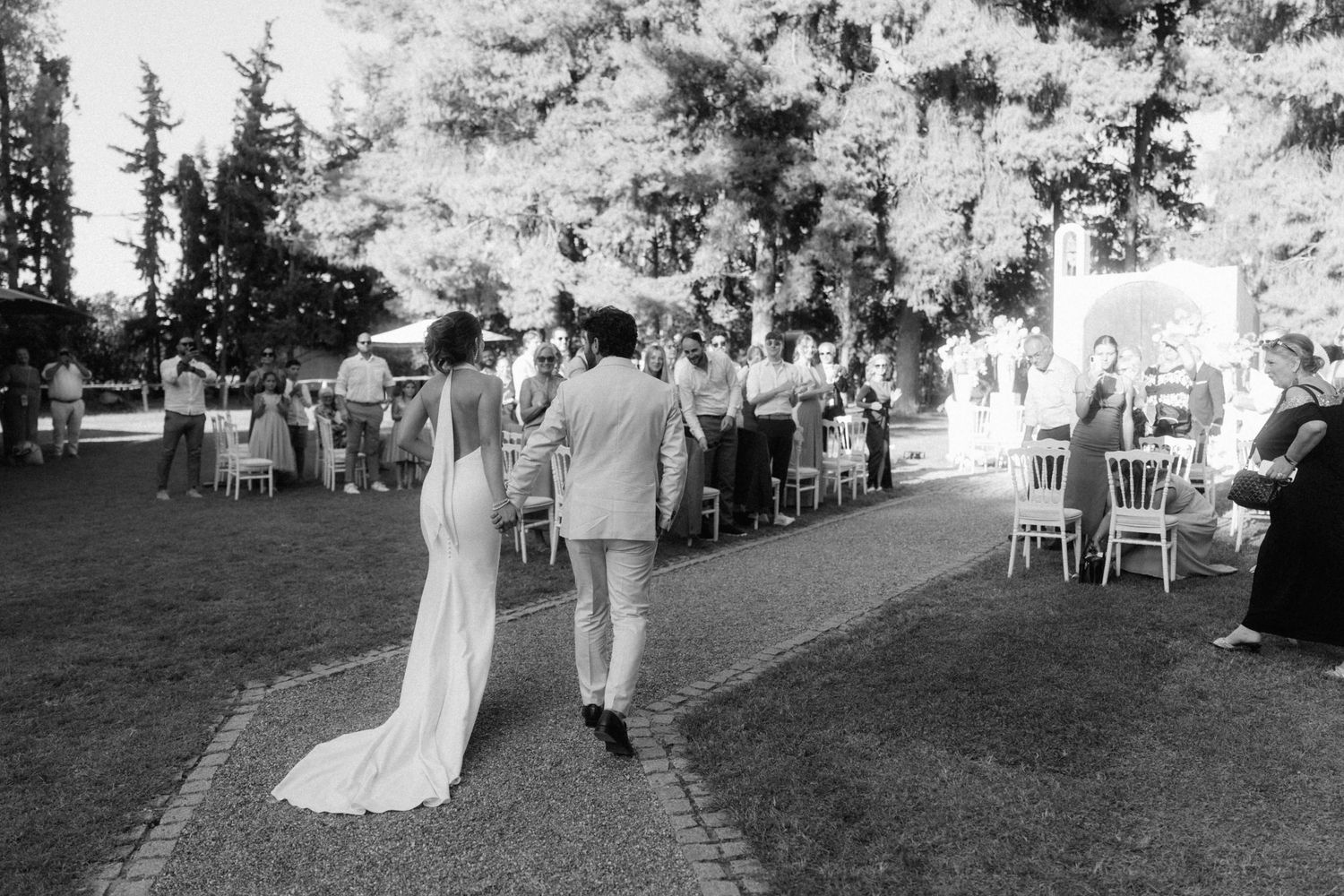 Black and white photo of bride and groom walking hand in hand toward wedding guests outdoors.