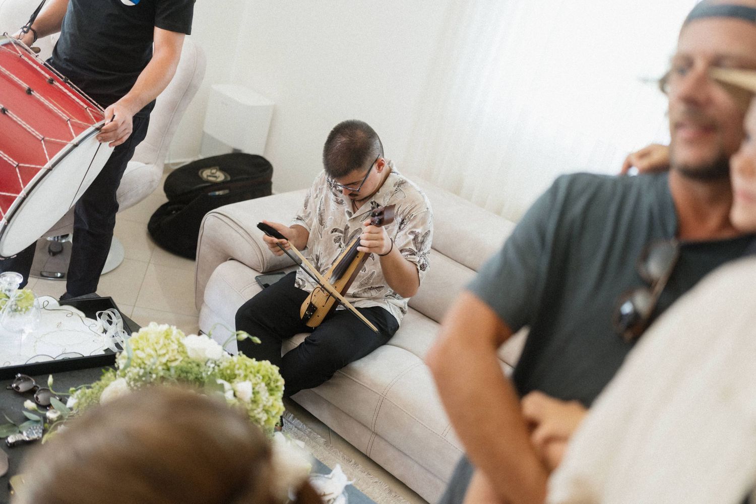 A child sits on a white sofa holding a violin while adults with instruments gather in a bright living room.