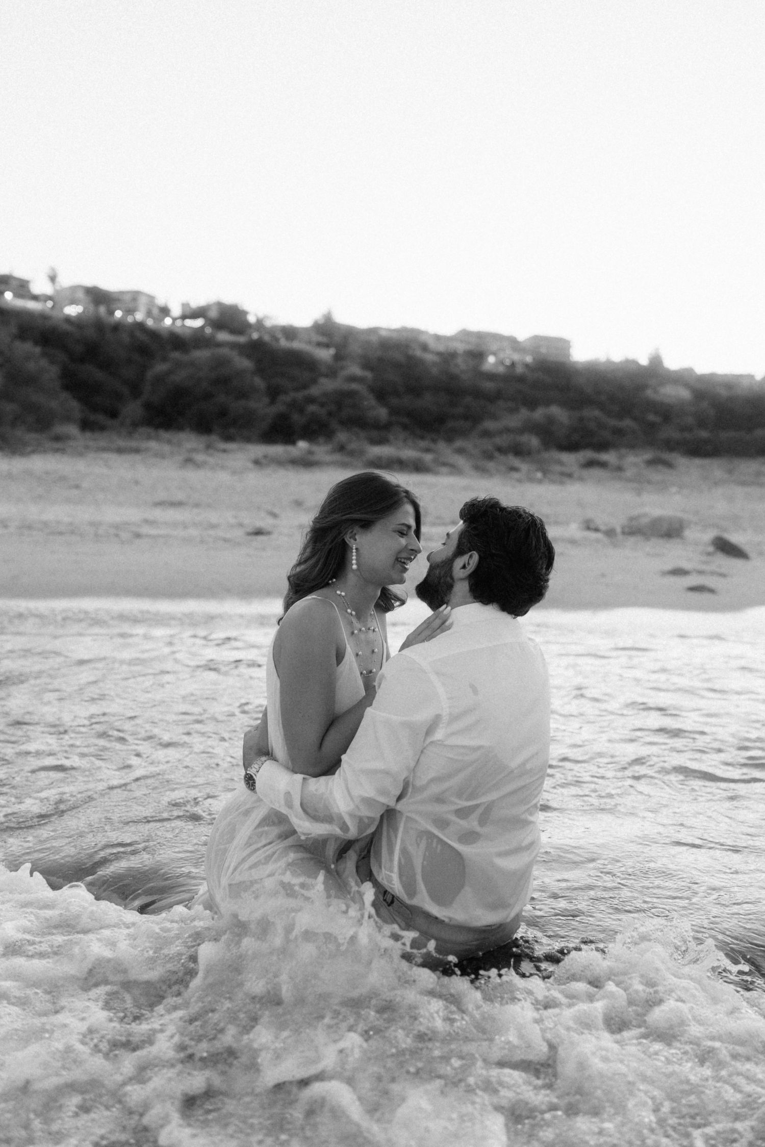 Black and white photo of a couple embracing in ocean waves on a beach, sharing a romantic moment in wet clothing.