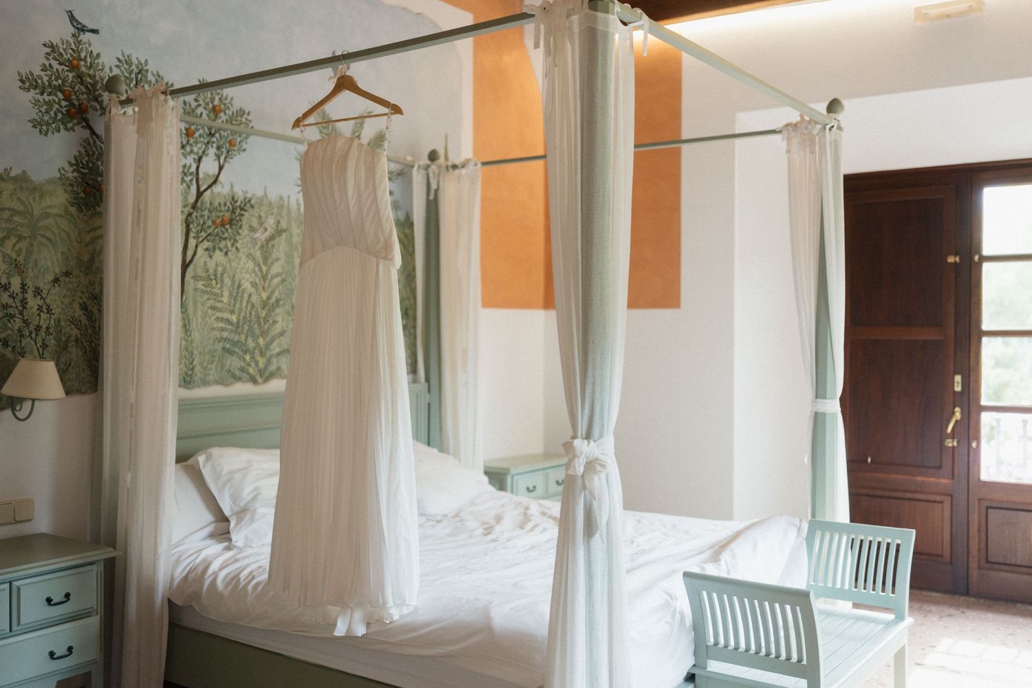 White wedding dress hanging from a canopy bed with sheer white curtains in a bright bedroom with a botanical mural and wooden French doors.