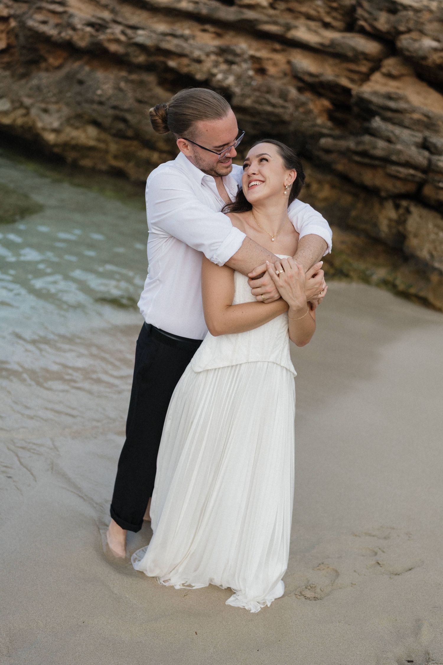 Smiling couple in white embracing on a sandy beach beside turquoise water and rocky cliffs during a seaside wedding photoshoot.