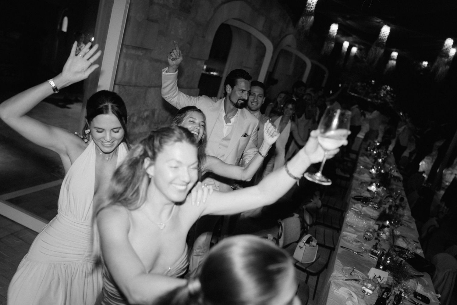 Black and white photo of joyful wedding guests dancing and raising glasses beside a long reception dinner table.