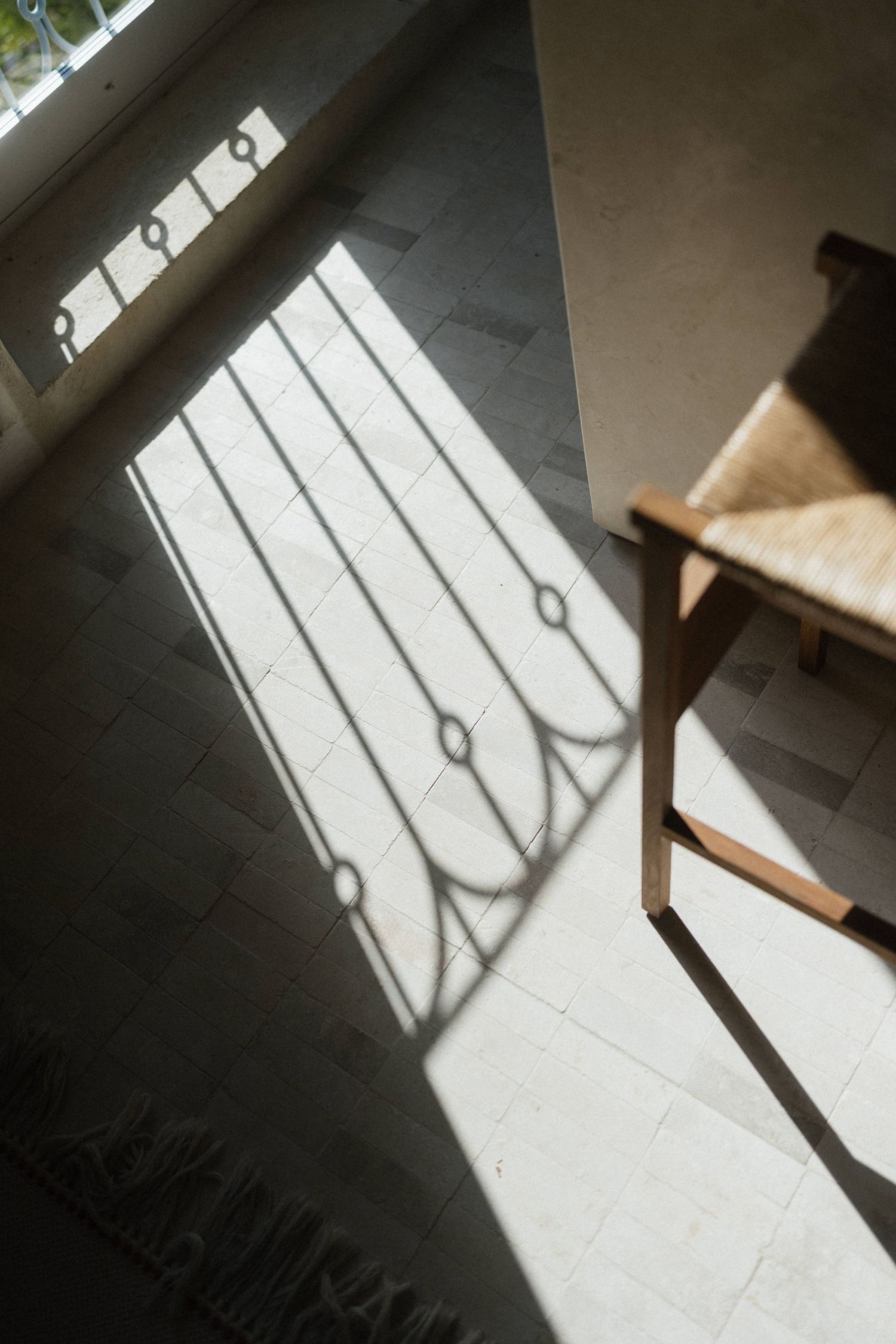 Sunlight casts ornate window shadows across a stone tile floor beside a wooden chair with a woven rush seat.