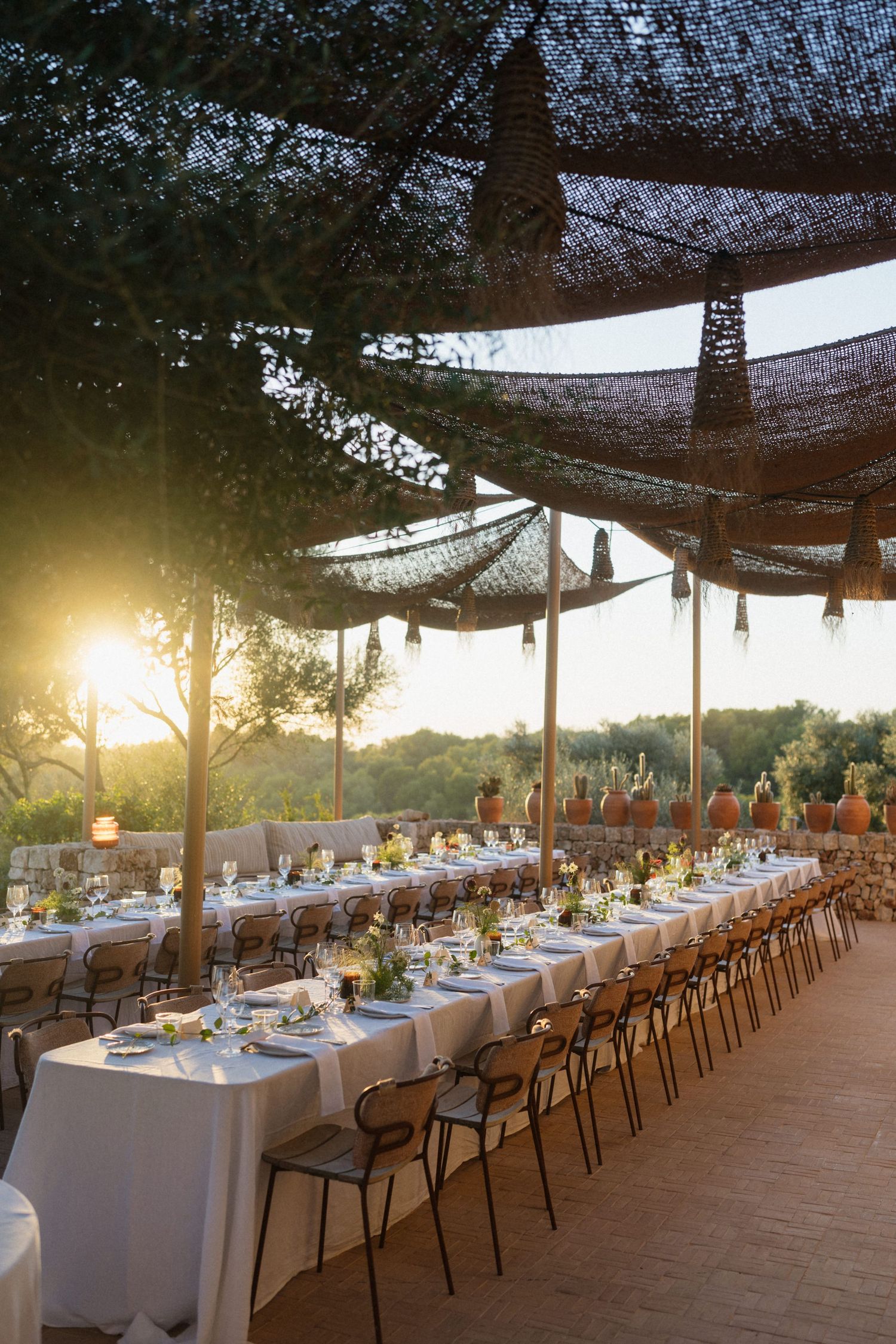 A long wedding banquet table with white linens and floral centerpieces under a woven canopy, glowing in golden sunset light.
