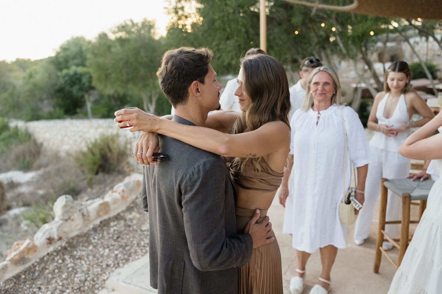 Couple embracing in a warm hug at an outdoor gathering while guests in white linen outfits smile and watch in a sunlit garden setting.