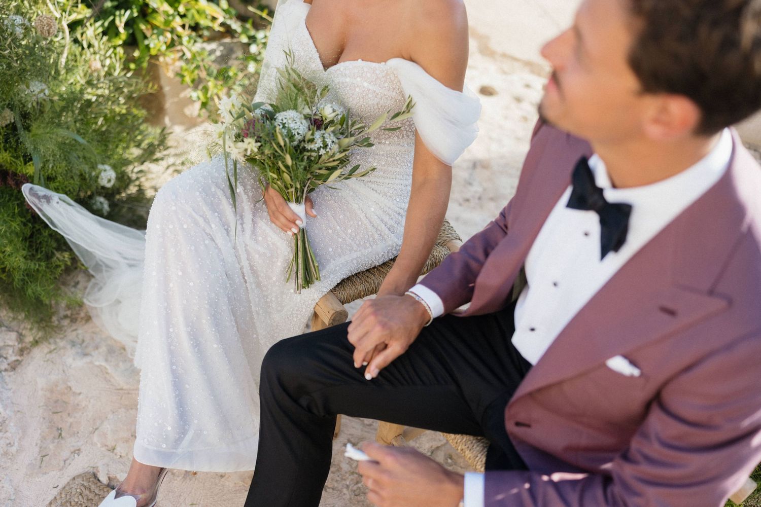 Bride in a white gown holding a bouquet sits beside a groom in a burgundy blazer and black bow tie on a sunny stone terrace.