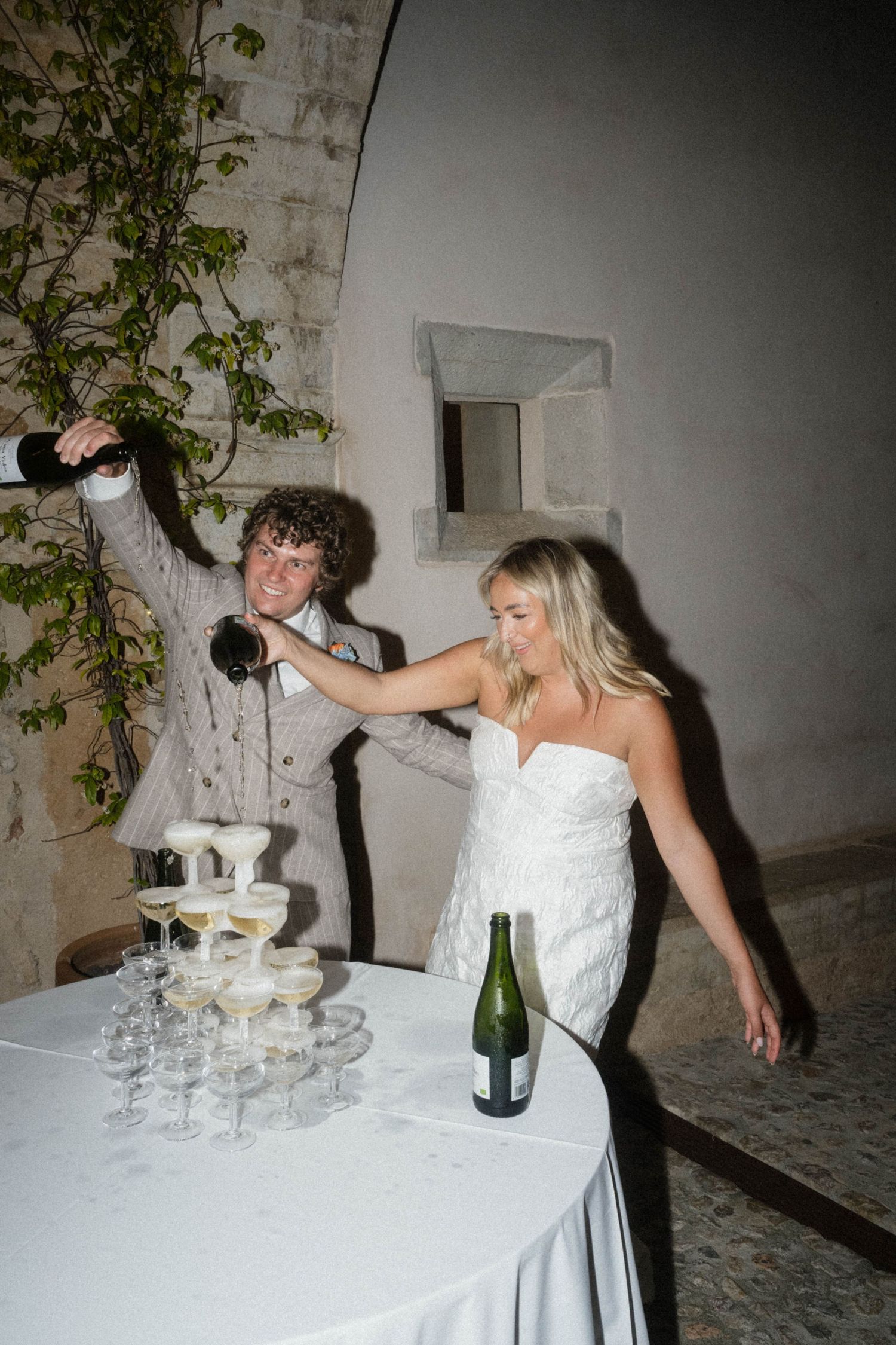 Bride in white gown and groom pour champagne tower at elegant wedding reception outdoors.