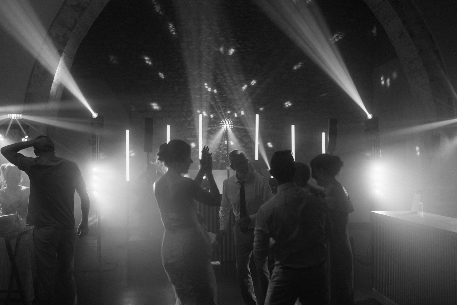 Black and white photo of people dancing in a nightclub with dramatic stage lighting and fog effects.