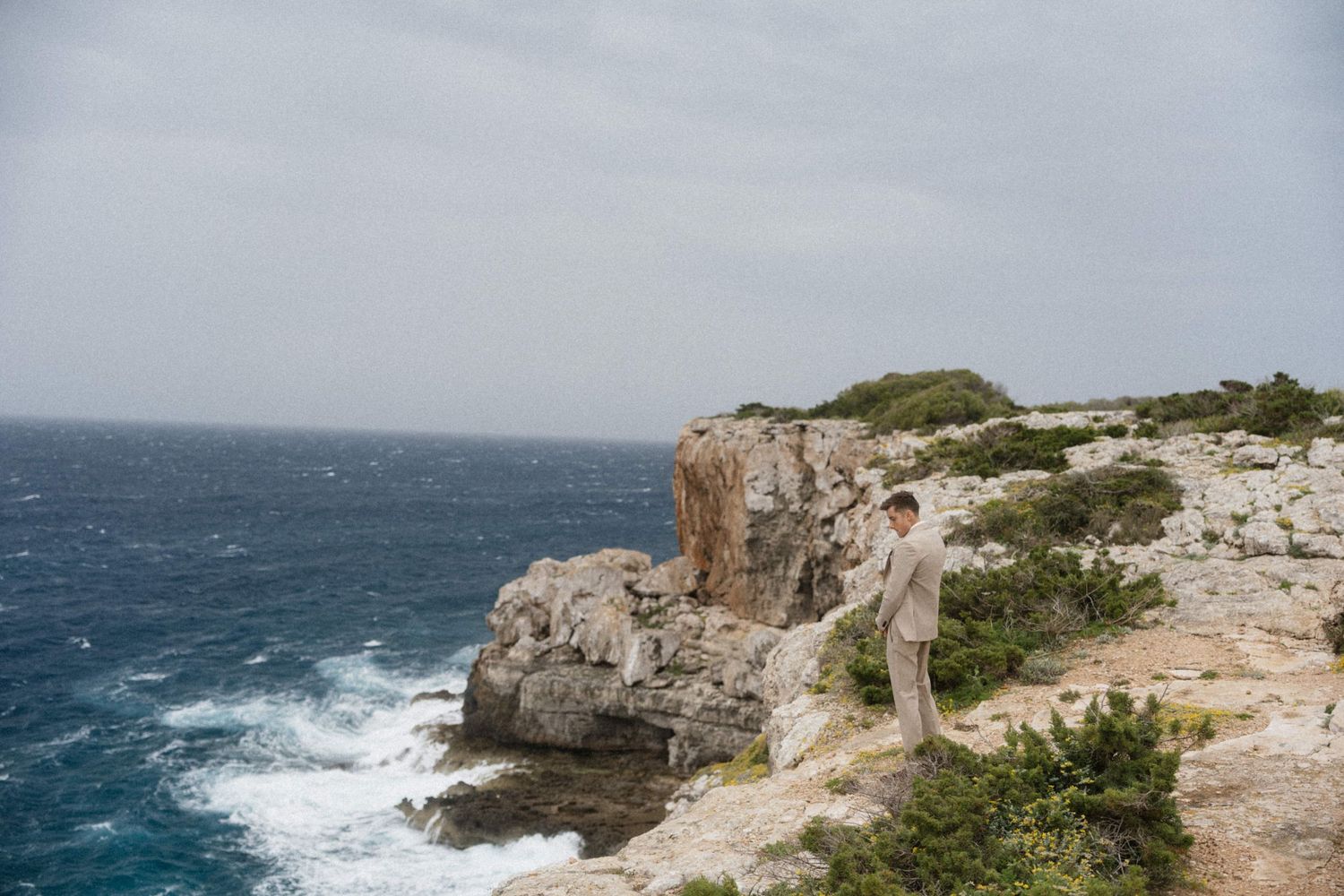Man in beige suit standing on rocky coastal cliffs overlooking turquoise ocean waves under overcast sky.