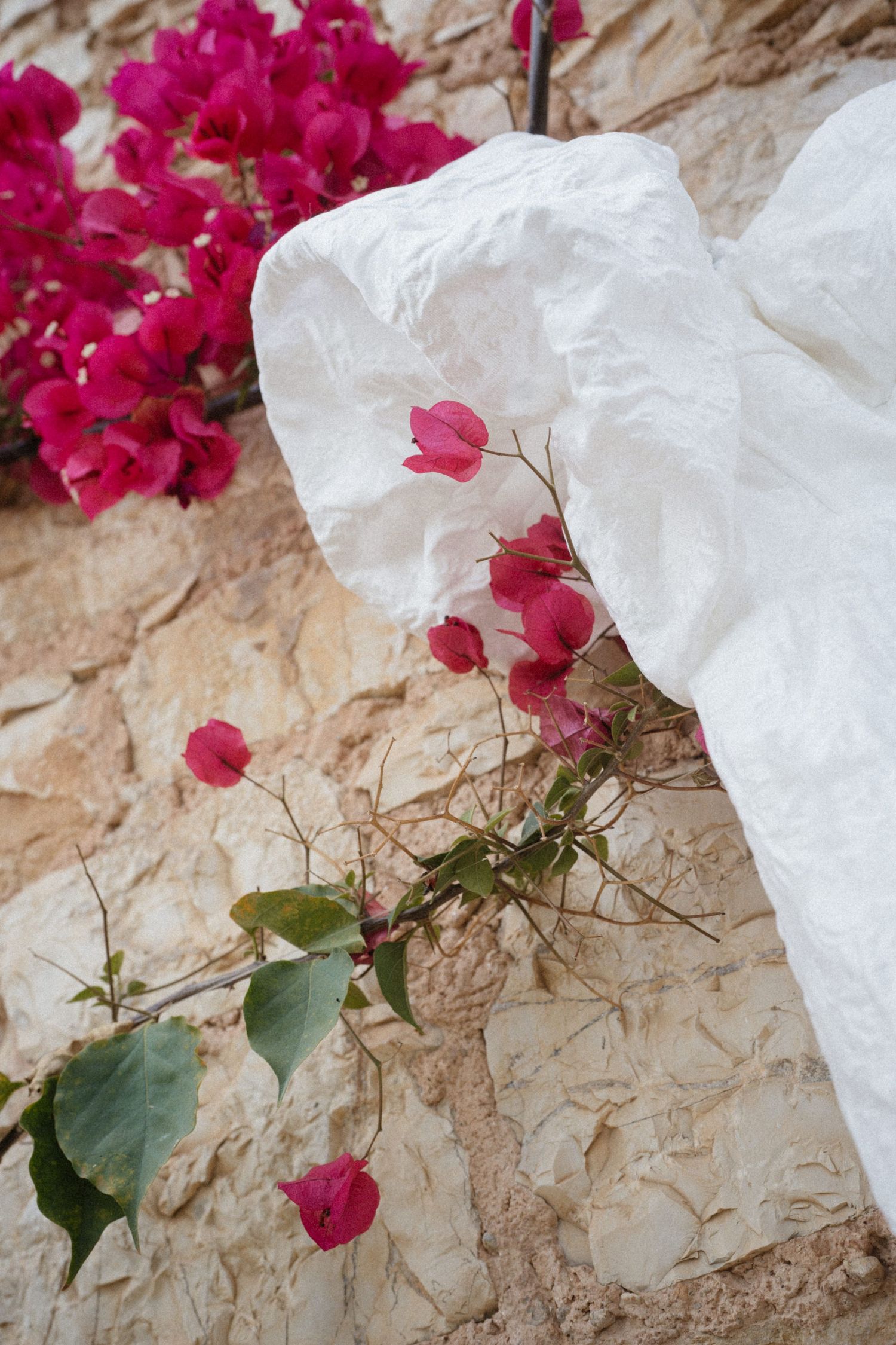 White wedding dress draped against a rustic stone wall with vibrant pink bougainvillea flowers.