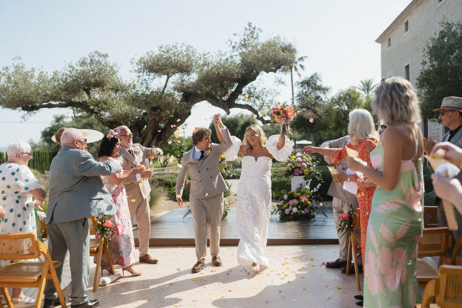 Joyful bride and groom walk down the aisle as guests toss petals at an outdoor wedding ceremony.