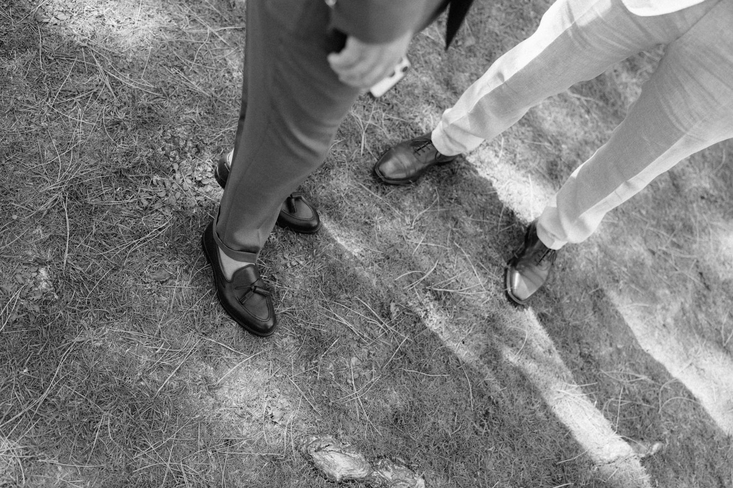 Black and white photo of two men in dress shoes and formal trousers standing on rustic ground at a wedding.