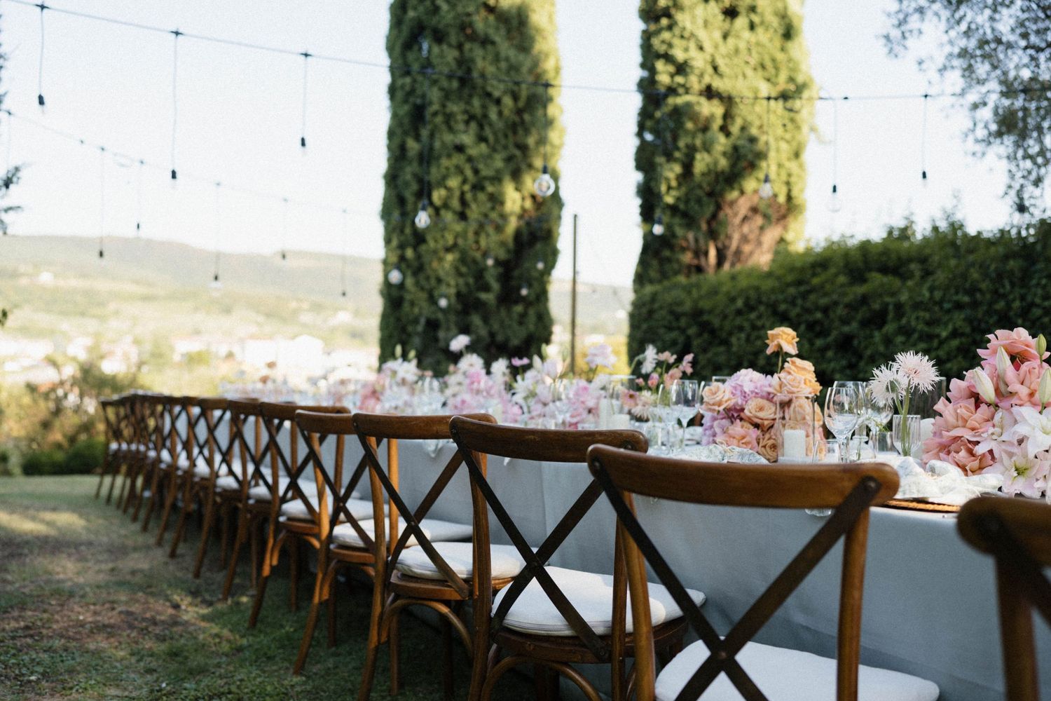 Elegant outdoor wedding reception with long wooden tables, floral centerpieces, string lights, and cypress trees in Tuscany.