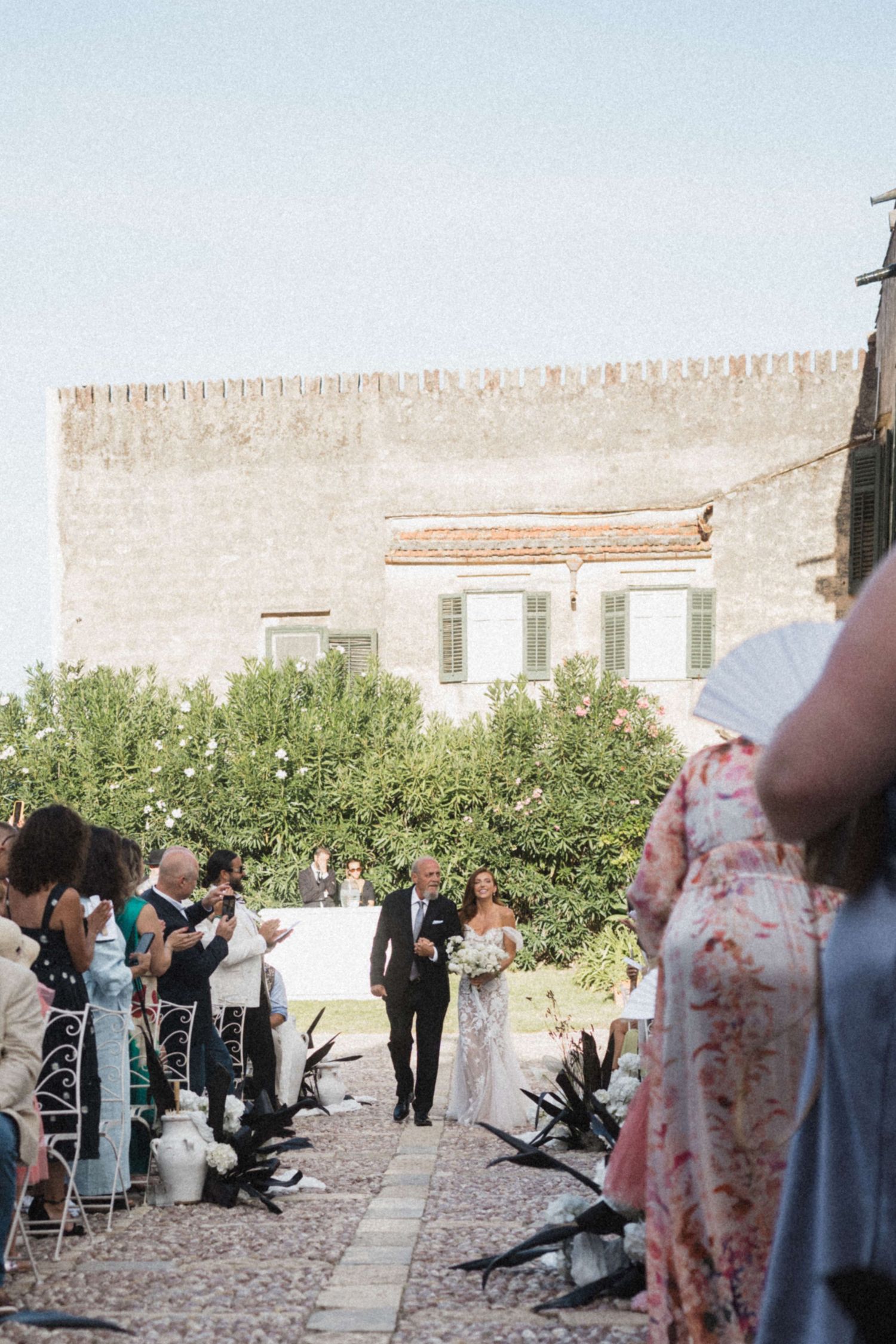 Bride walks down aisle with escort at outdoor Mediterranean wedding ceremony.