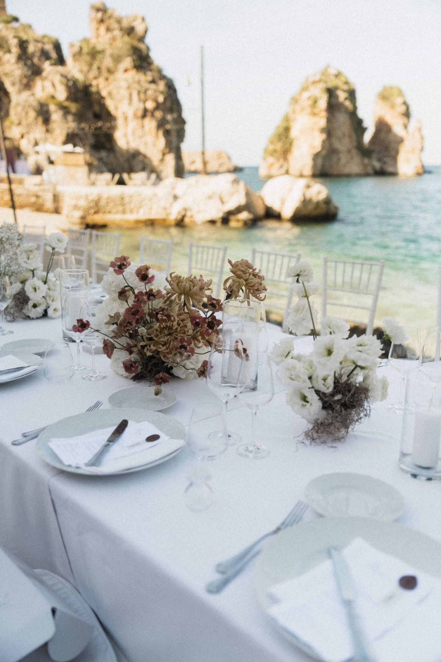 Stunning coastal wedding reception table with white floral centerpieces and dramatic sea stack rock backdrop.