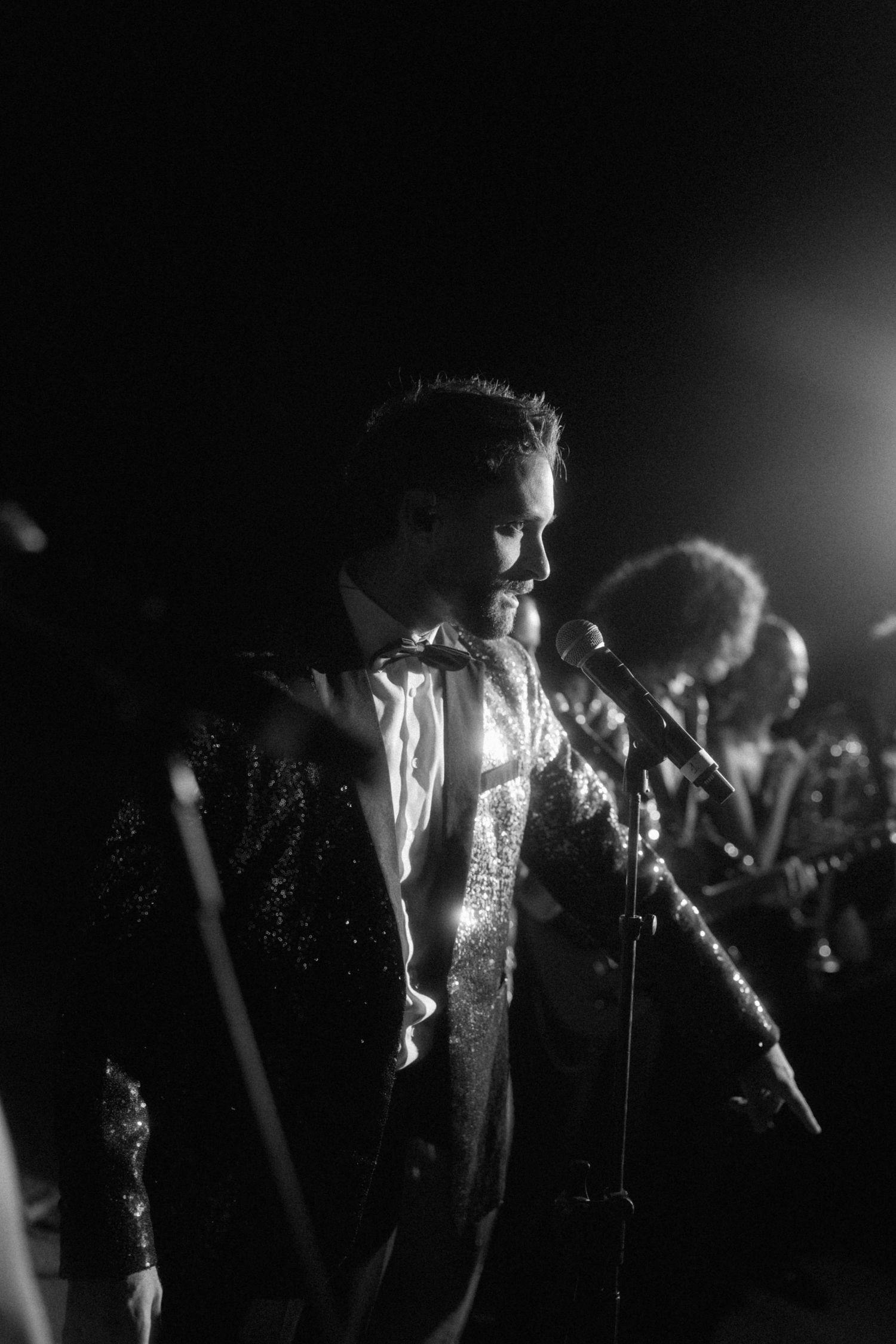 Black and white photo of male performer in sequin jacket singing into microphone at wedding reception.