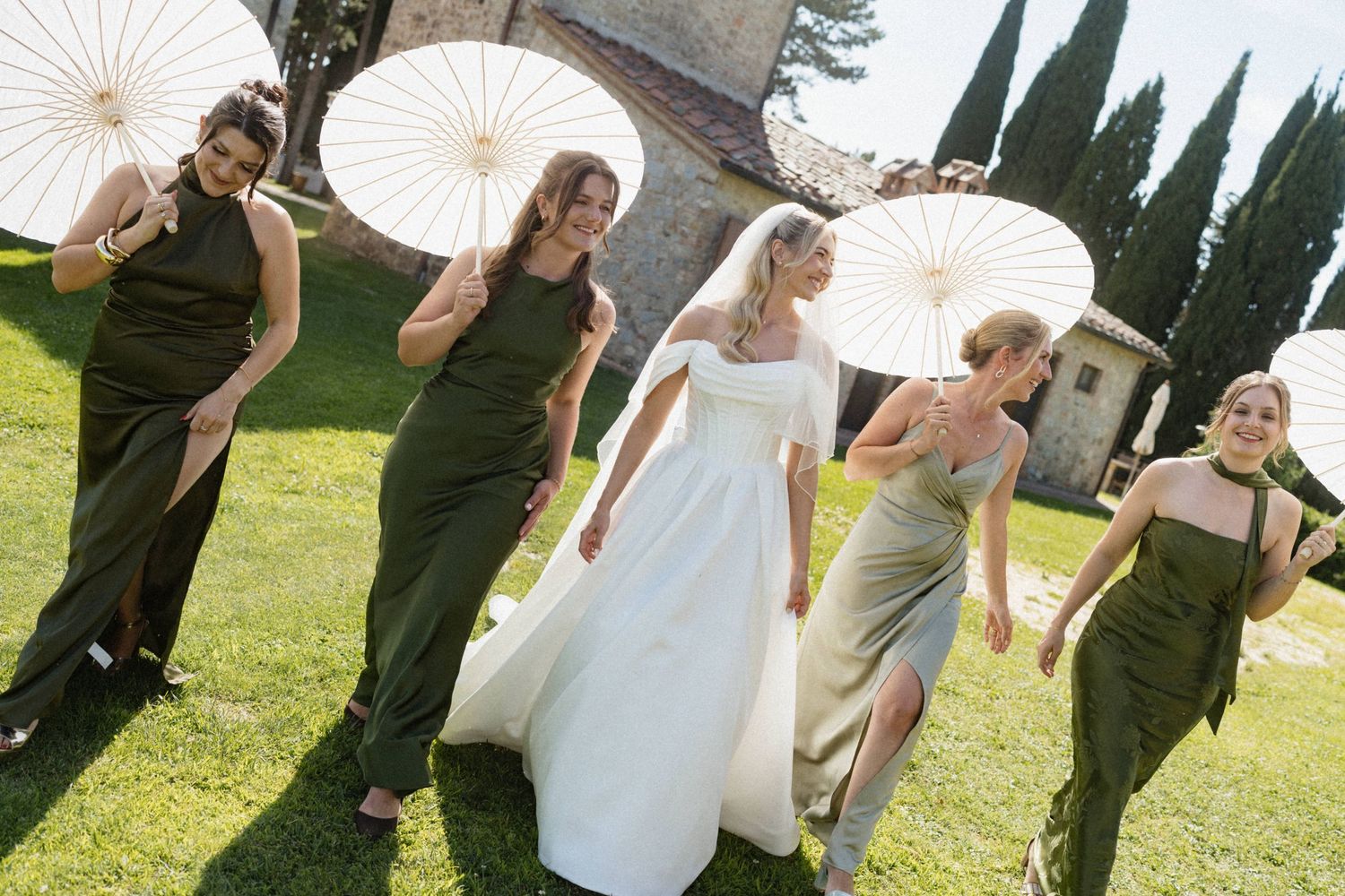Bride and bridesmaids in olive green dresses walking with white parasols on a sunny Tuscan wedding day.