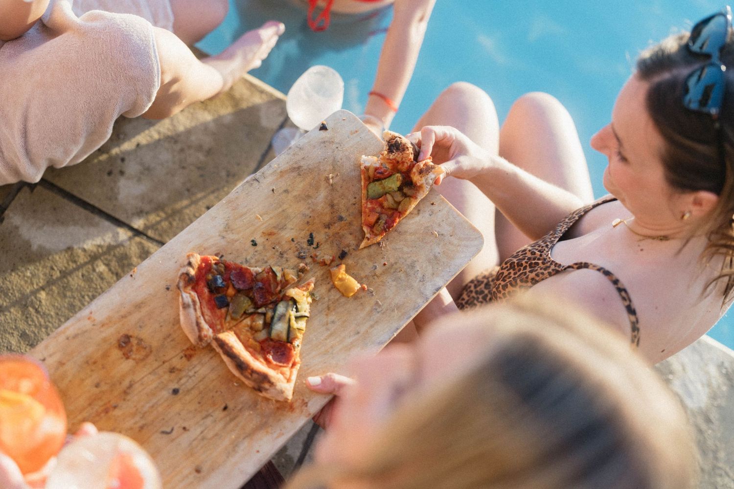 Women in swimsuits enjoying pizza slices served on a wooden board at a sunny poolside gathering.
