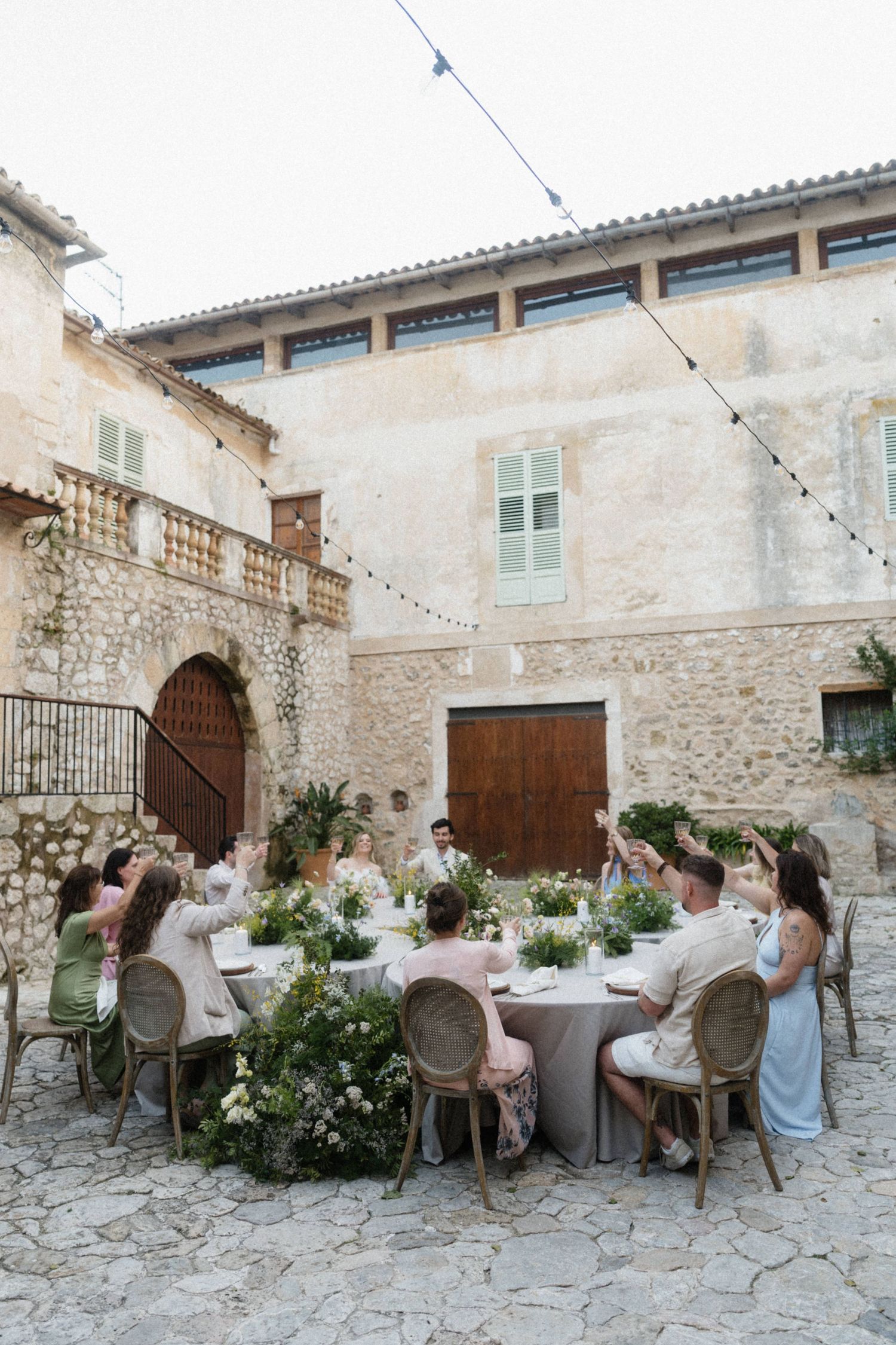Wedding guests seated at a round table in a rustic stone courtyard with string lights above.