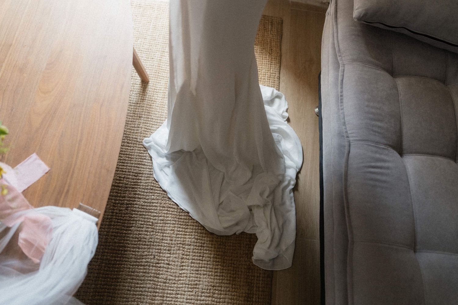 Close-up of a white wedding dress train pooling on a natural fiber rug beside a wooden table and gray sofa.